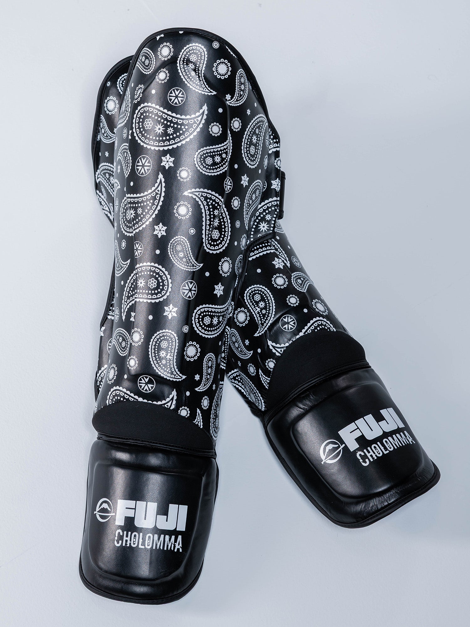 A pair of black CholoMMA Paisley Shin Guards with white paisley patterns are crossed on a light gray surface.