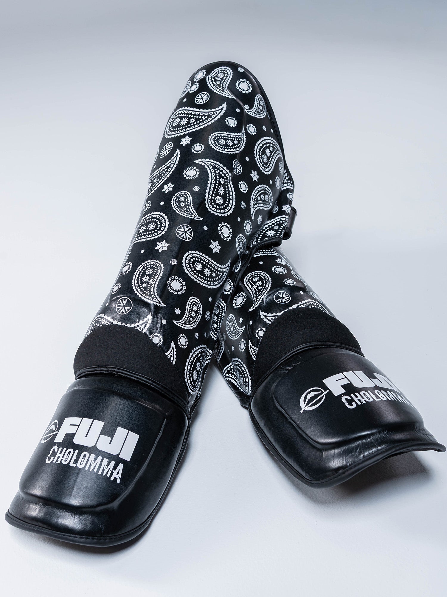 A pair of CholoMMA Paisley Shin Guards in black with white paisley patterns and FUJI CHOLONMA branding on the foot pads, displayed on a light gray surface.