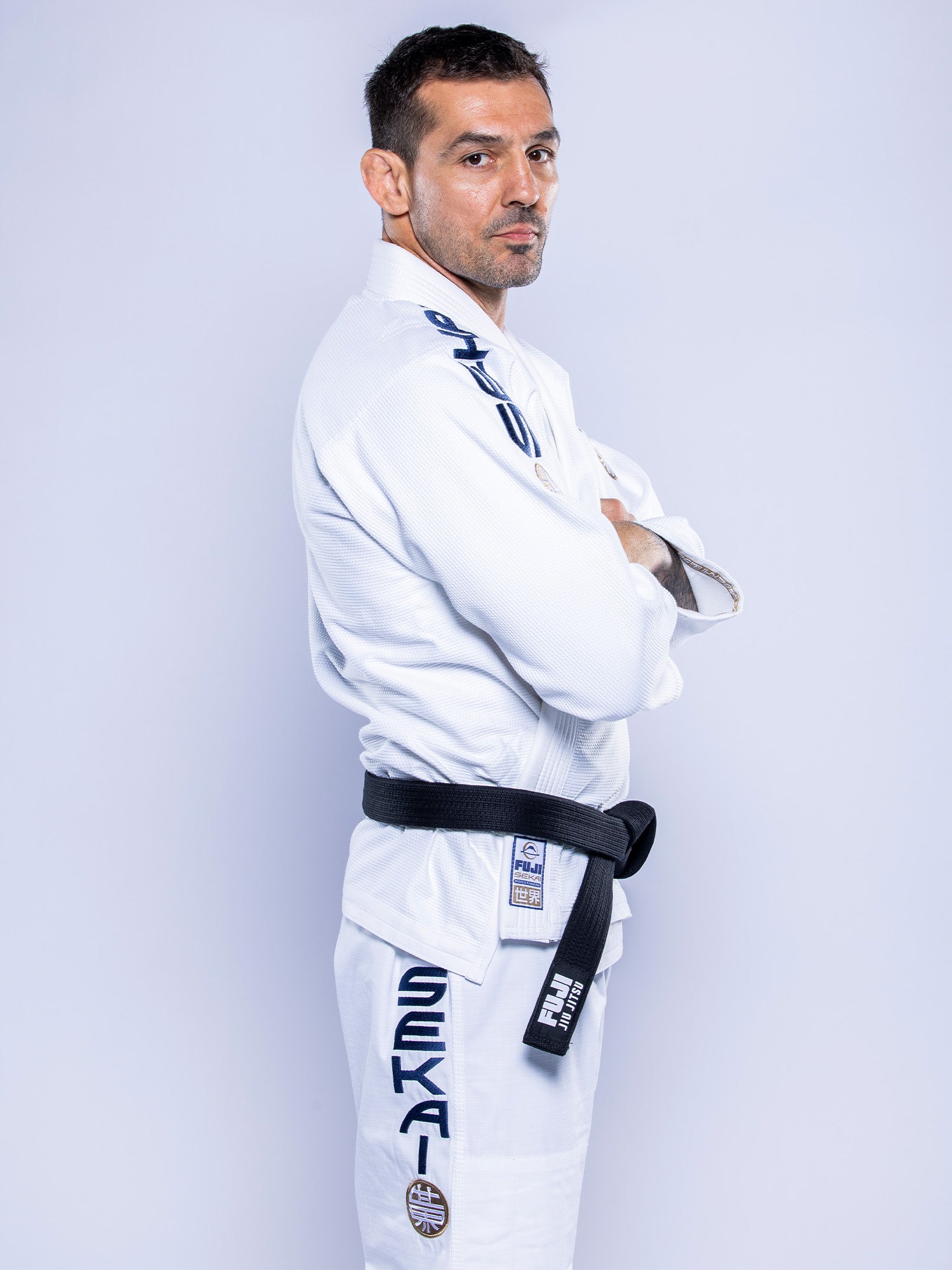A man wearing the SEKAI 3.0 BJJ Gi White Navy with blue and black lettering and patches, plus a black belt, stands sideways with arms crossed, looking at the camera against a plain light background.