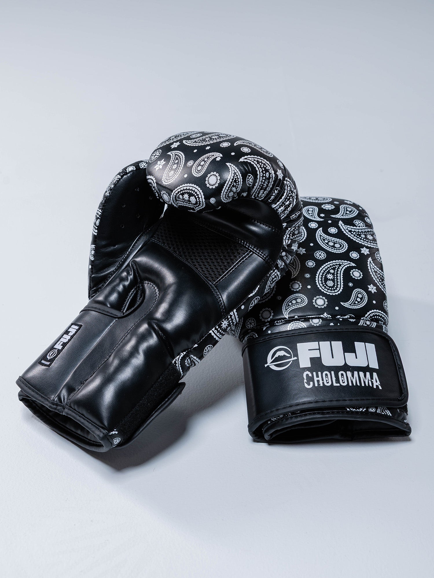 The CholoMMA Paisley 16oz Boxing Gloves feature a black design with white paisley patterns and FUJI CHOLOMMA branding on the wrist straps, displayed on a light gray surface.