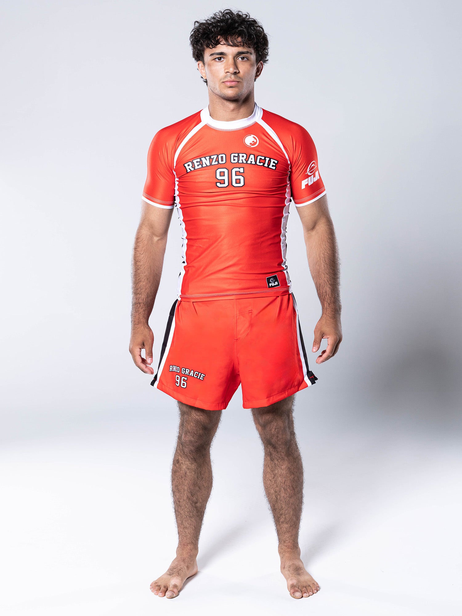 A man with curly hair stands barefoot, wearing the Renzo Gracie '96 Jersey Short Sleeve Rashguard in red and matching shorts, both featuring “Renzo Gracie 96” text, posing against a plain light background.
