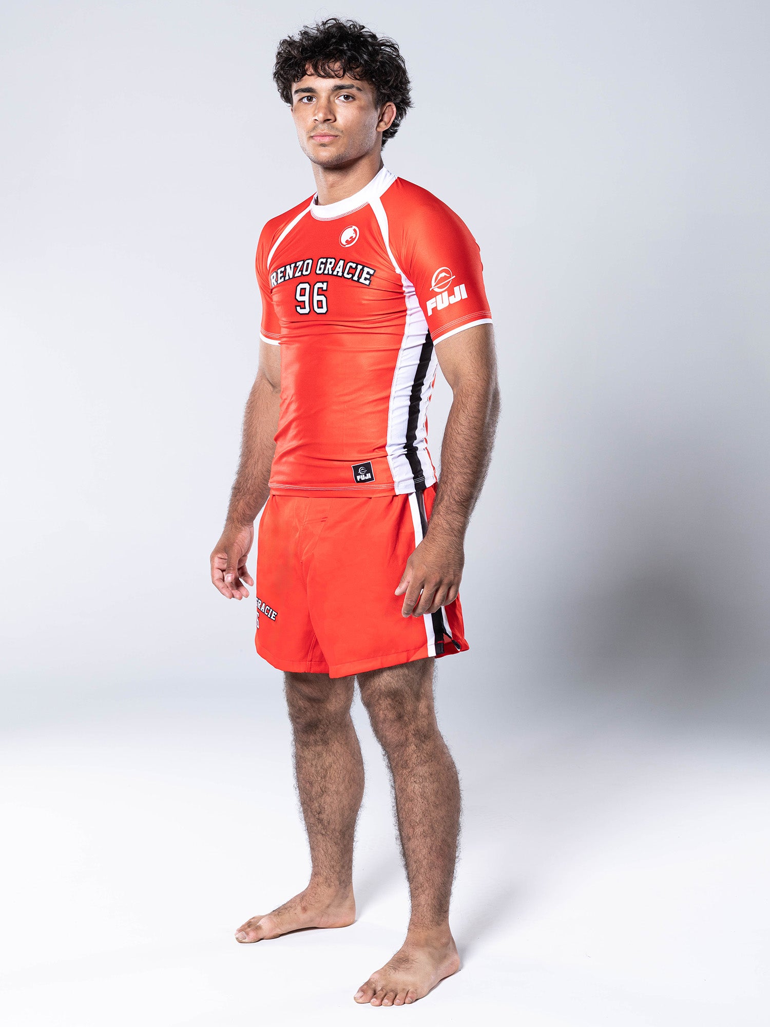 A young man stands barefoot wearing a Renzo Gracie '96 Jersey Short Sleeve Rashguard in red with matching shorts, posing against a plain white background.