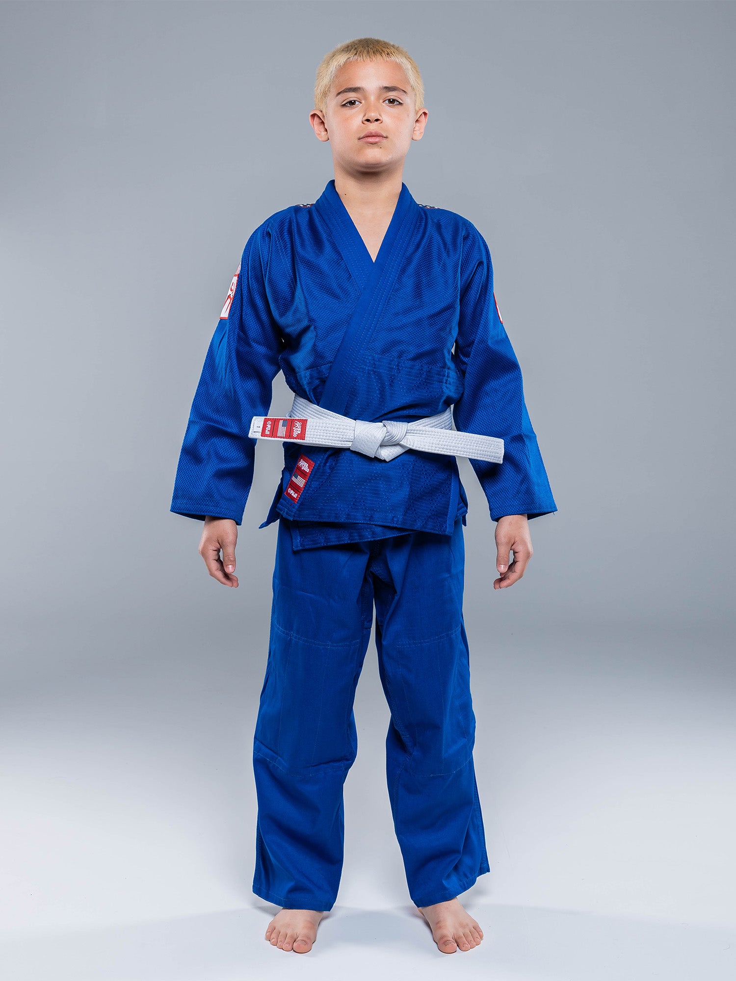 A young boy stands barefoot against a gray background, wearing the USA Judo Single Weave Gi 2.0 Blue with a white belt tied at the waist. He looks straight ahead with a neutral expression.