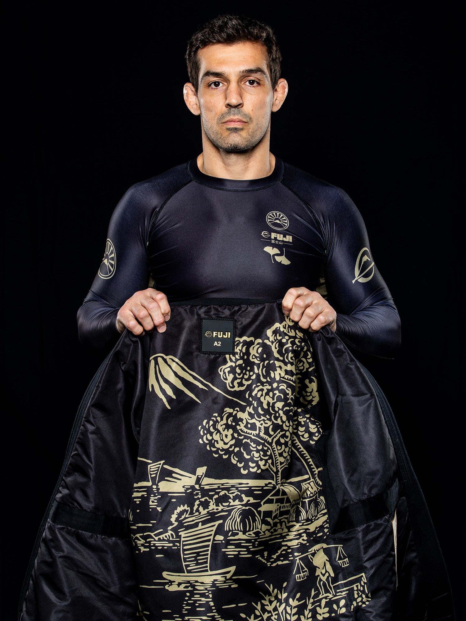 A man in a dark athletic outfit stands against a black background, holding open the Heritage Keisho Lightweight BJJ Gi Black to reveal its gold Japanese-style landscape lining.