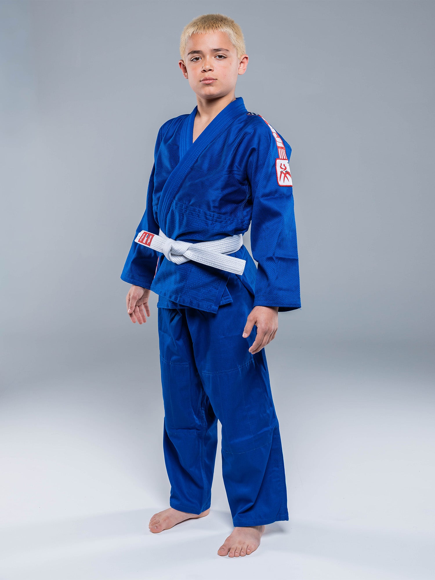 A young boy with short blond hair stands barefoot on a gray background, wearing the USA Judo Single Weave Gi 2.0 Blue with a white belt tied around his waist.