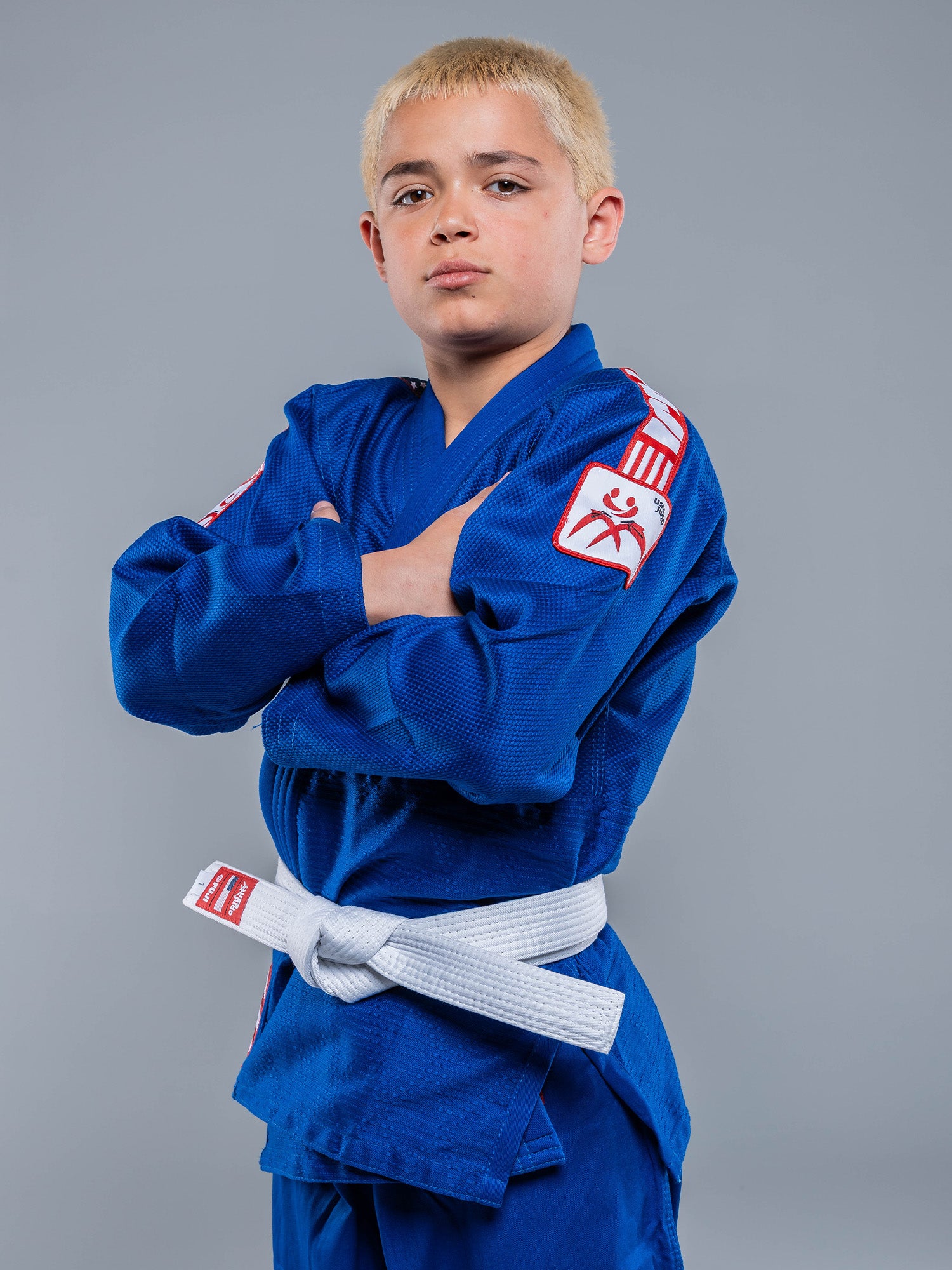 A young boy with short blond hair stands confidently with arms crossed, wearing the USA Judo Single Weave Gi 2.0 Blue and a white belt against a plain gray background.