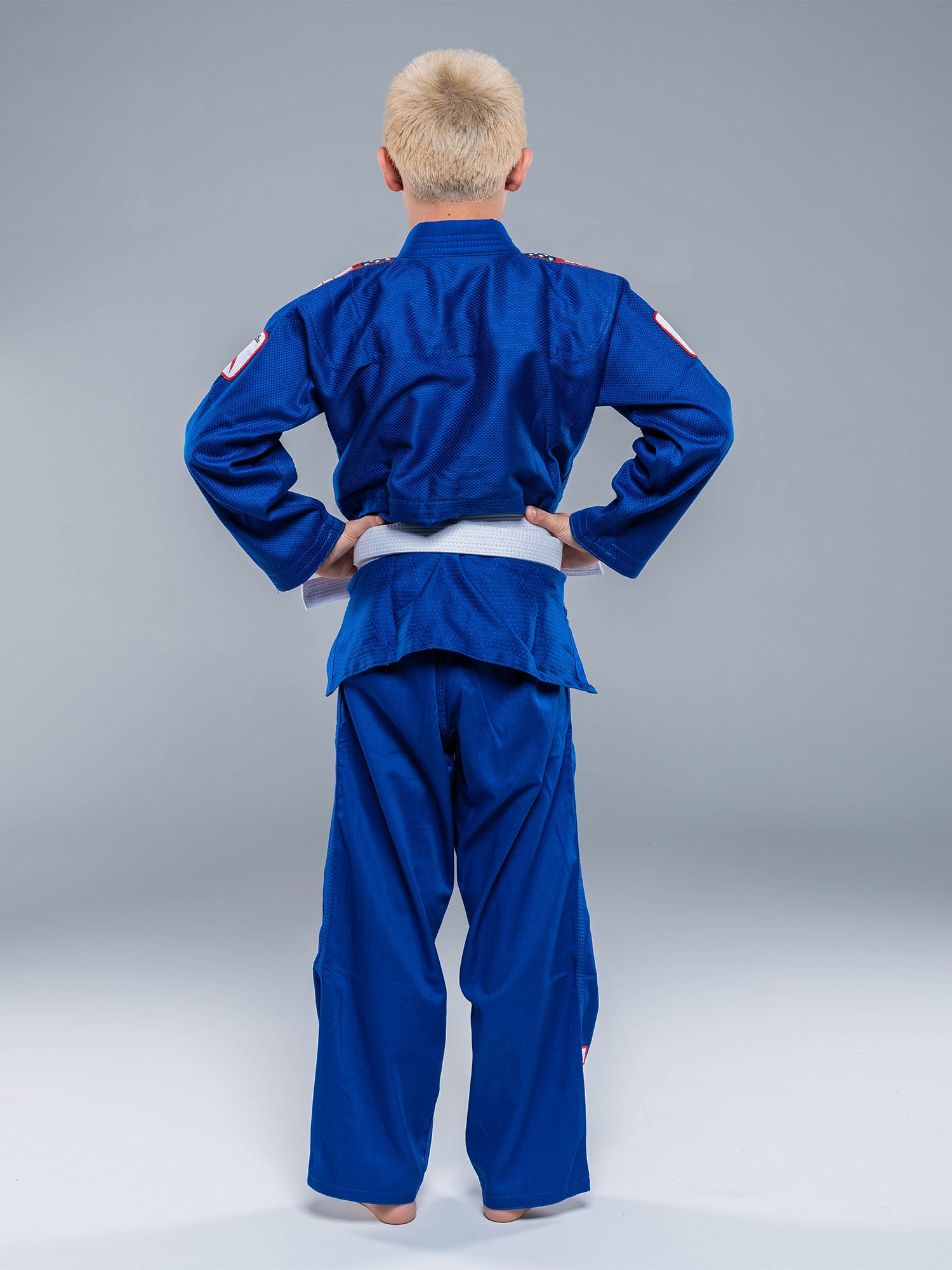 A barefoot child with short blond hair, facing away from the camera, wears a USA Judo Single Weave Gi 2.0 Blue and a white belt against a plain gray background.