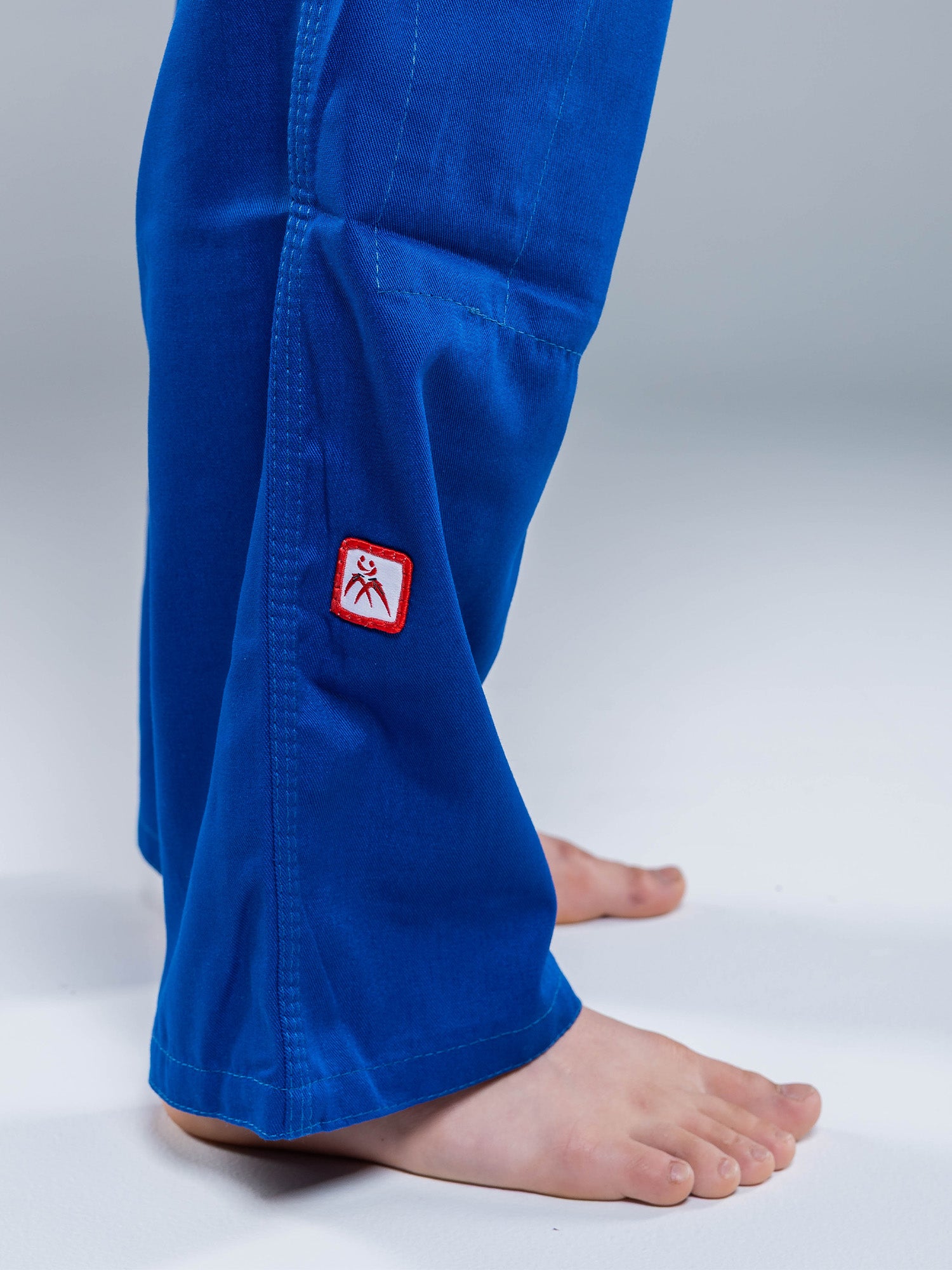 A close-up of bare feet and lower legs in the USA Judo Single Weave Gi 2.0 Blue, featuring a small red and white embroidered patch near the hem against a plain, neutral background.