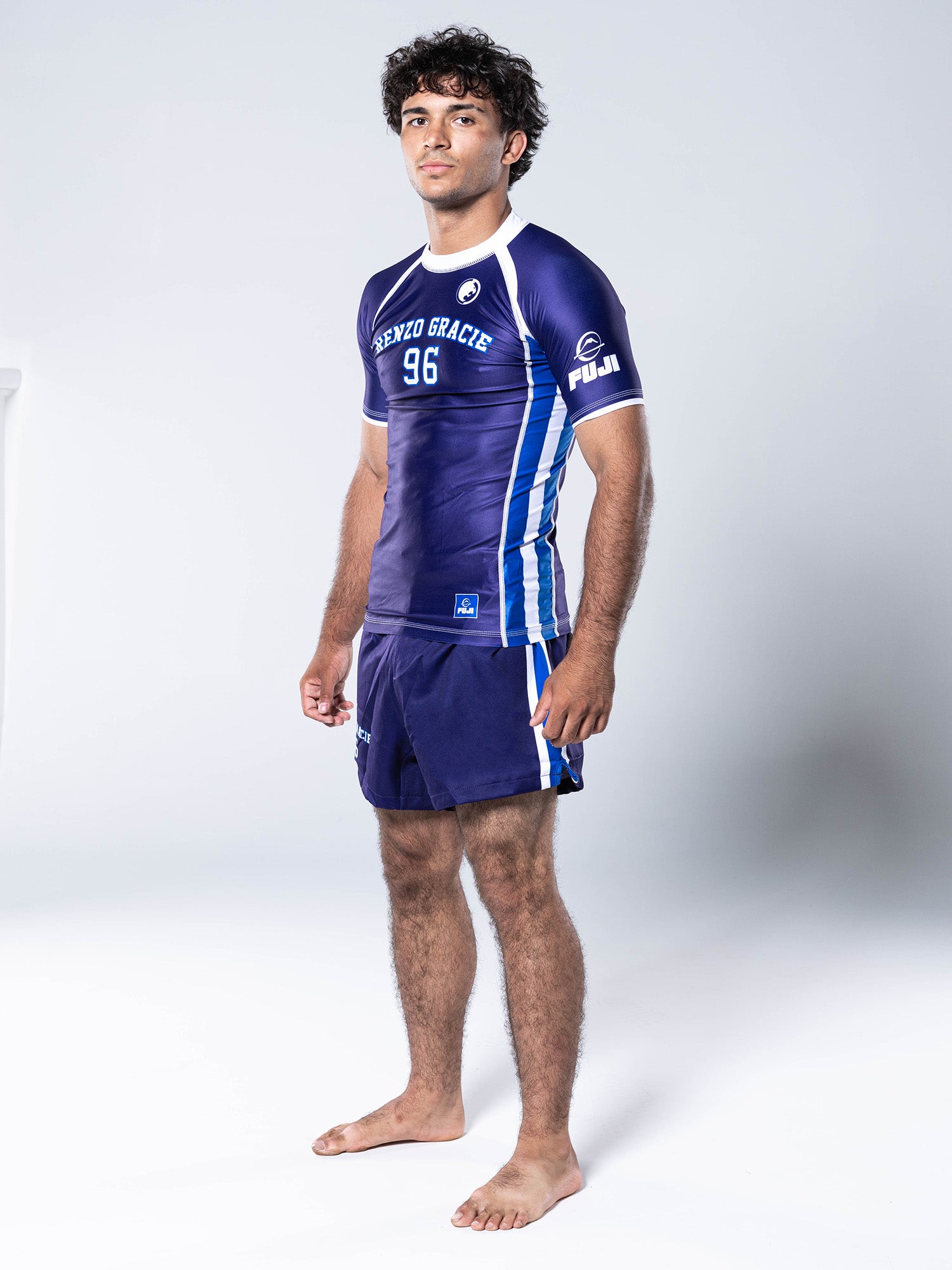 A man stands barefoot on a white background, wearing the Renzo Gracie '96 Jersey Short Sleeve Rashguard Blue and matching shorts, looking toward the camera.