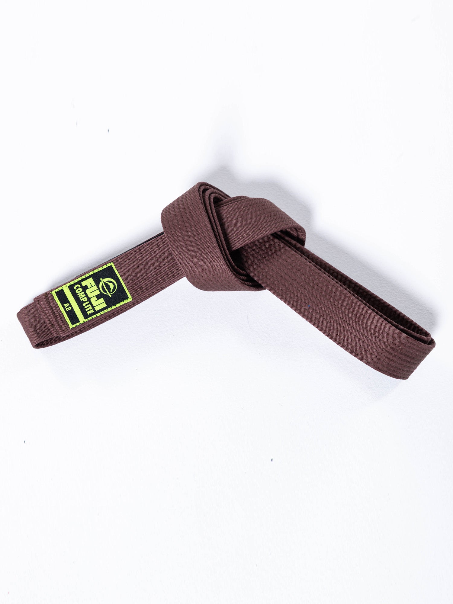 The BJJ Comp Lite Belt Brown, featuring a textured pattern, is loosely tied in a knot on a white background. A green and yellow tag with text is attached to one end of the belt.
