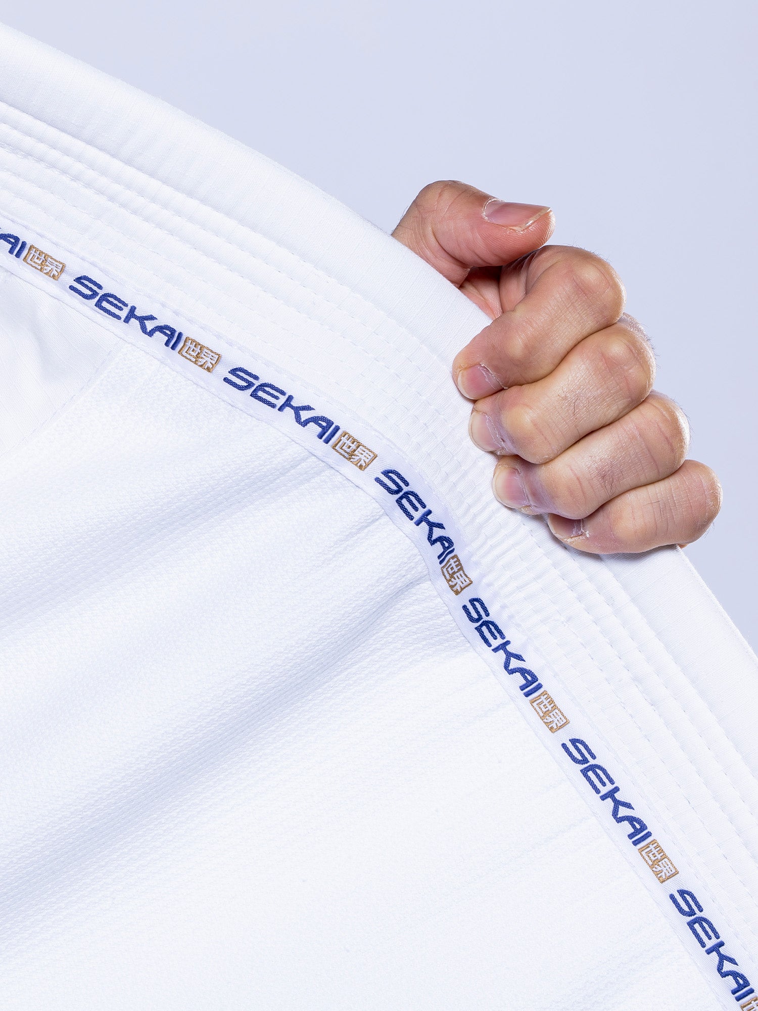 A hand grips the collar of the SEKAI 3.0 BJJ Gi White Navy, featuring "SEKAI" repeated in blue along the trim. The background is plain and light-colored.