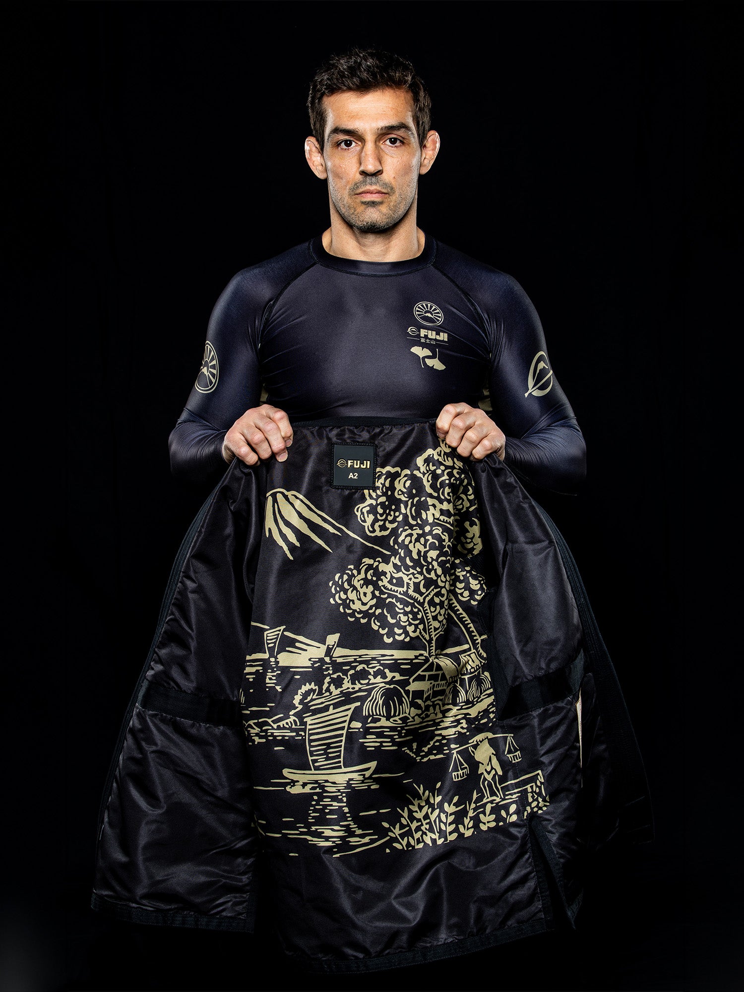 A man in a dark athletic outfit holds open the black jacket from the Heritage Keishō Edition Bundle, revealing an intricate gold illustration of a landscape with a boat, trees, and mountains inside the lining.