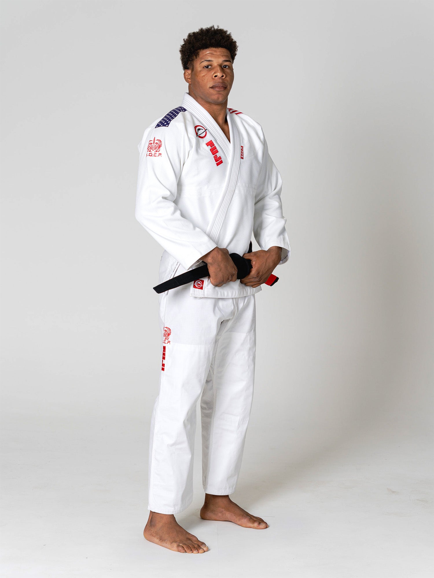 A man wearing the S.O.C.P. Sekai BJJ Gi 2.0 White with red and blue patches stands barefoot on a white background, holding a black belt tied around his waist.