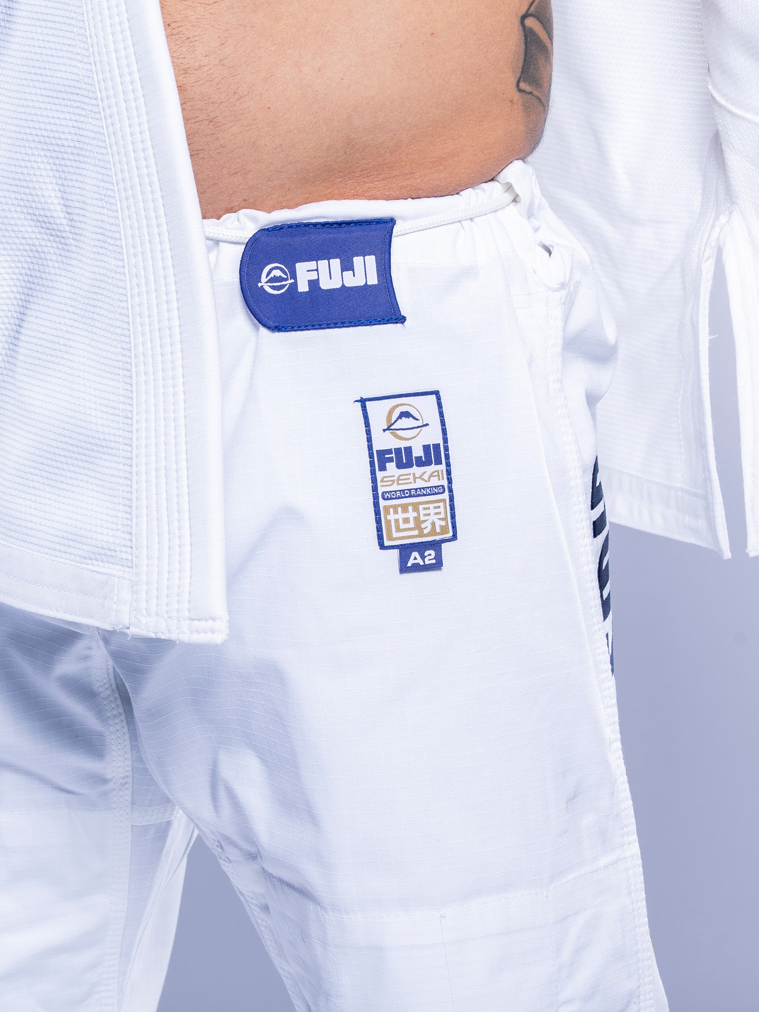 Close-up of a person wearing white SEKAI 3.0 BJJ Gi pants with navy patches and embroidery, featuring the FUJI logo and Japanese characters, against a light background.