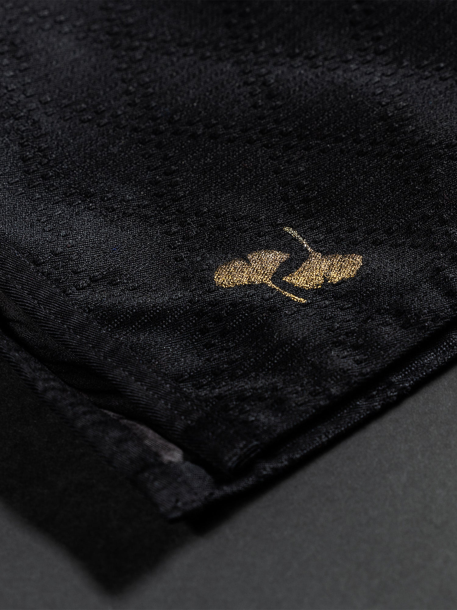 Close-up of the Womens Heritage Keisho Lightweight BJJ Gi in black, showcasing its textured fabric and a small, gold embroidered logo featuring a pair of high-heeled shoes.