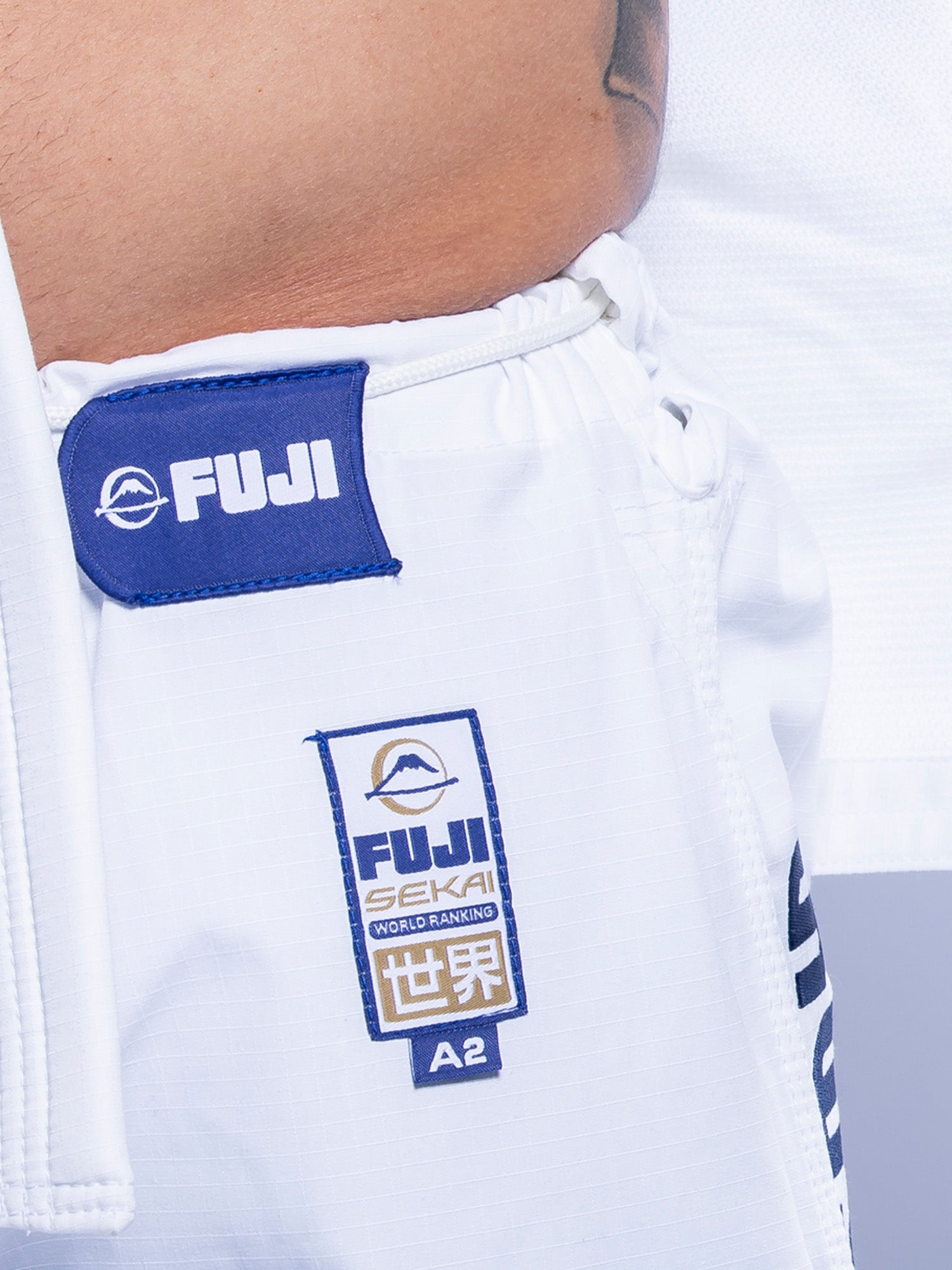 Close-up of a person wearing the SEKAI 3.0 BJJ Gi White Navy pants with a blue Fuji patch and FUJI SEKAI WORLD RANKING label featuring Japanese characters; part of a bare torso and gi jacket is also visible.