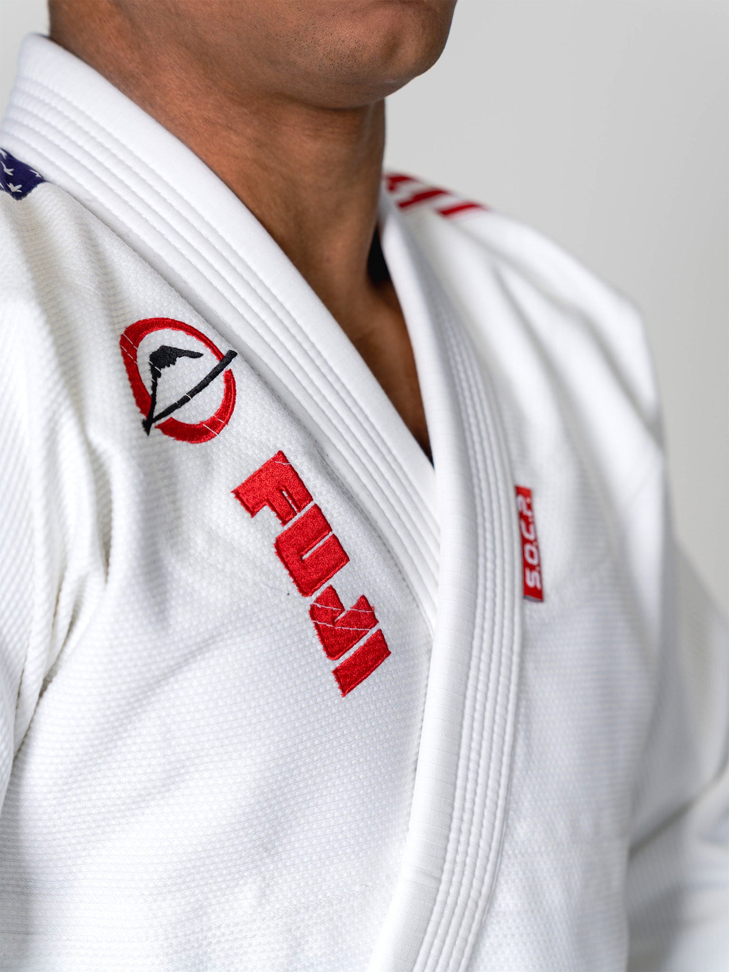 A person models the S.O.C.P. Sekai BJJ Gi 2.0 White, featuring red FUJI and logo patches on the chest and shoulder, photographed from shoulders to mid-torso against a plain background.