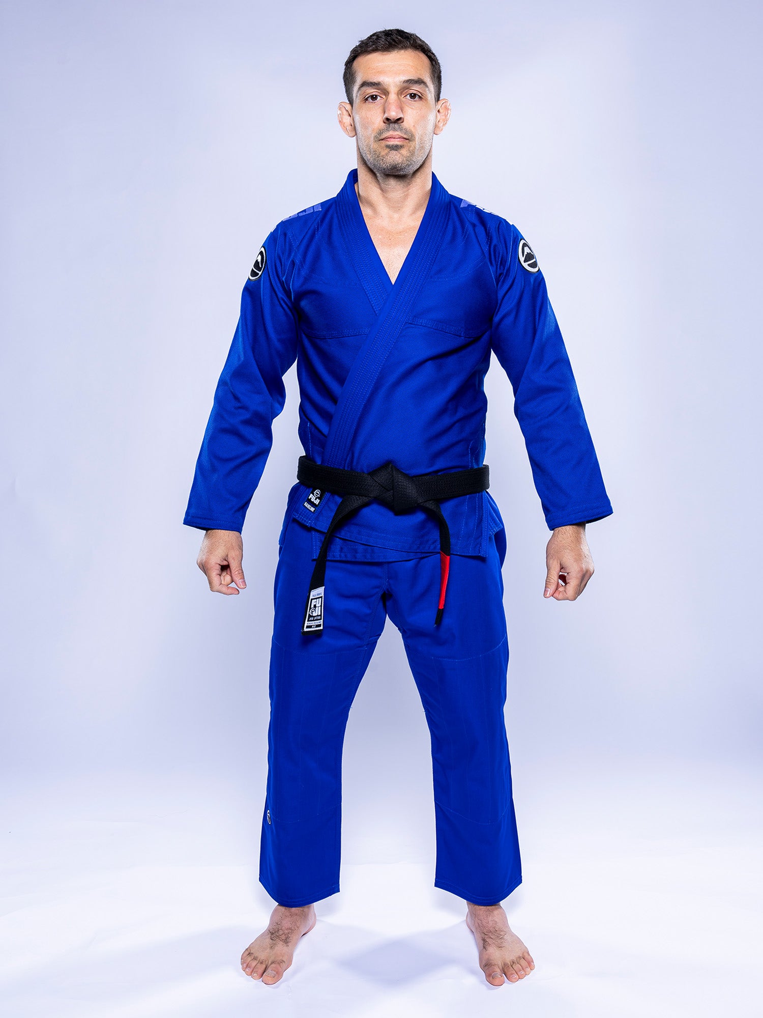 A man stands barefoot on a white background, facing forward with relaxed arms, wearing the Baseline 2.0 BJJ Gi Blue White and a black belt.