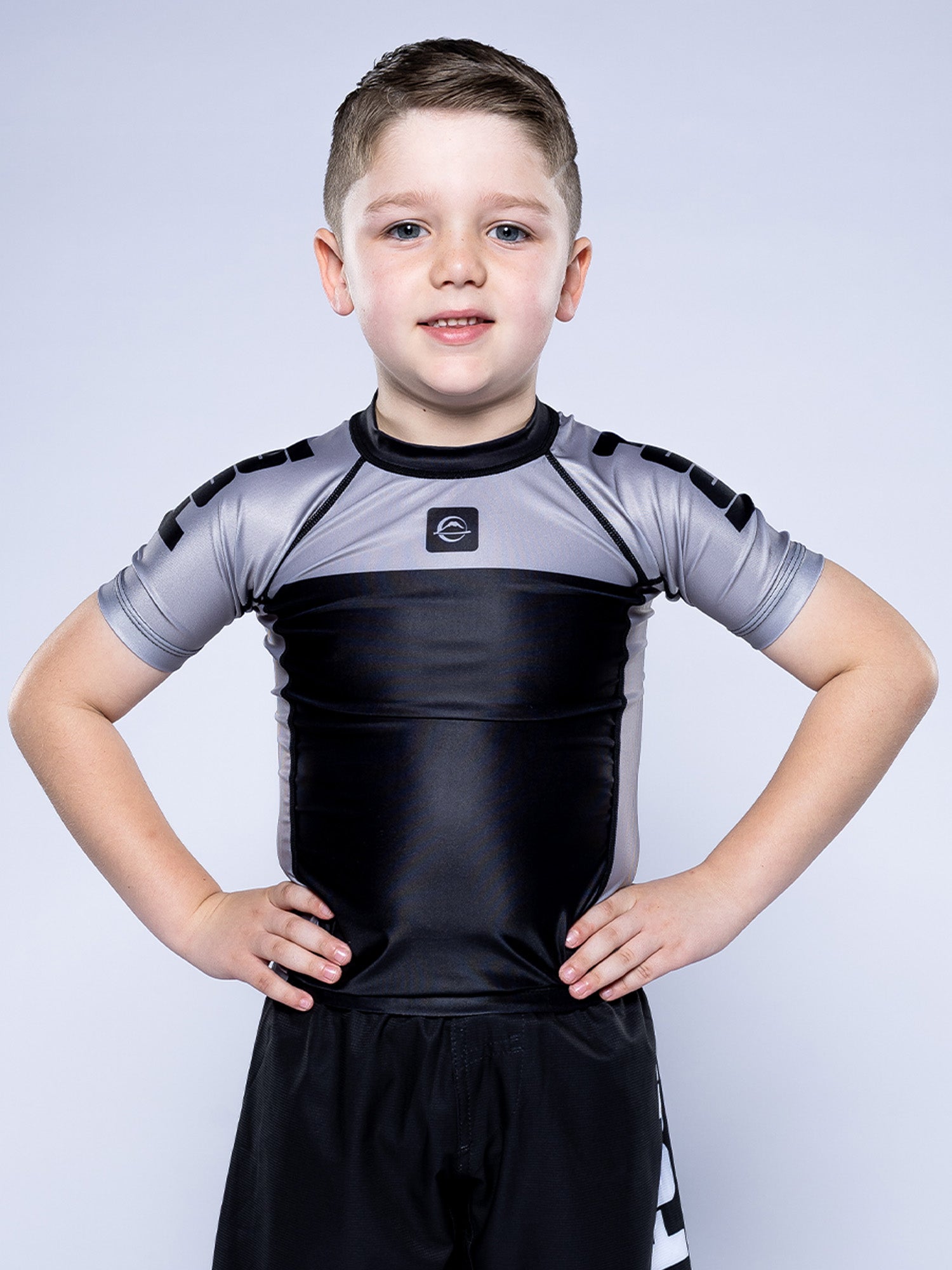 A young boy stands confidently with hands on hips, wearing the Kids Competition Ranked Rashguard Grey and matching shorts, smiling at the camera against a plain light background.