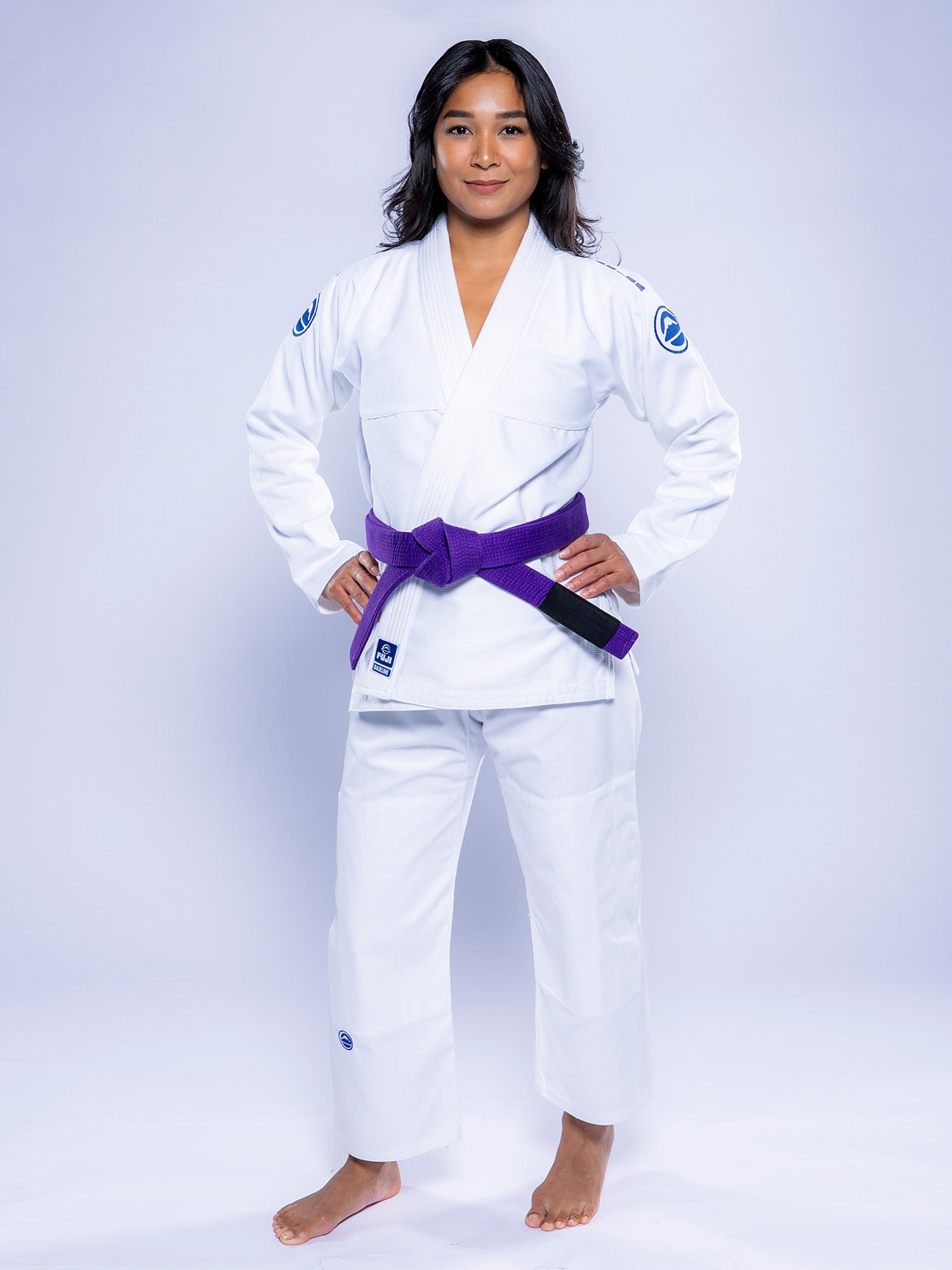 A woman stands confidently with hands on hips, barefoot, wearing the Womens Baseline 2.0 BJJ Gi White Navy and a purple belt against a plain white background.