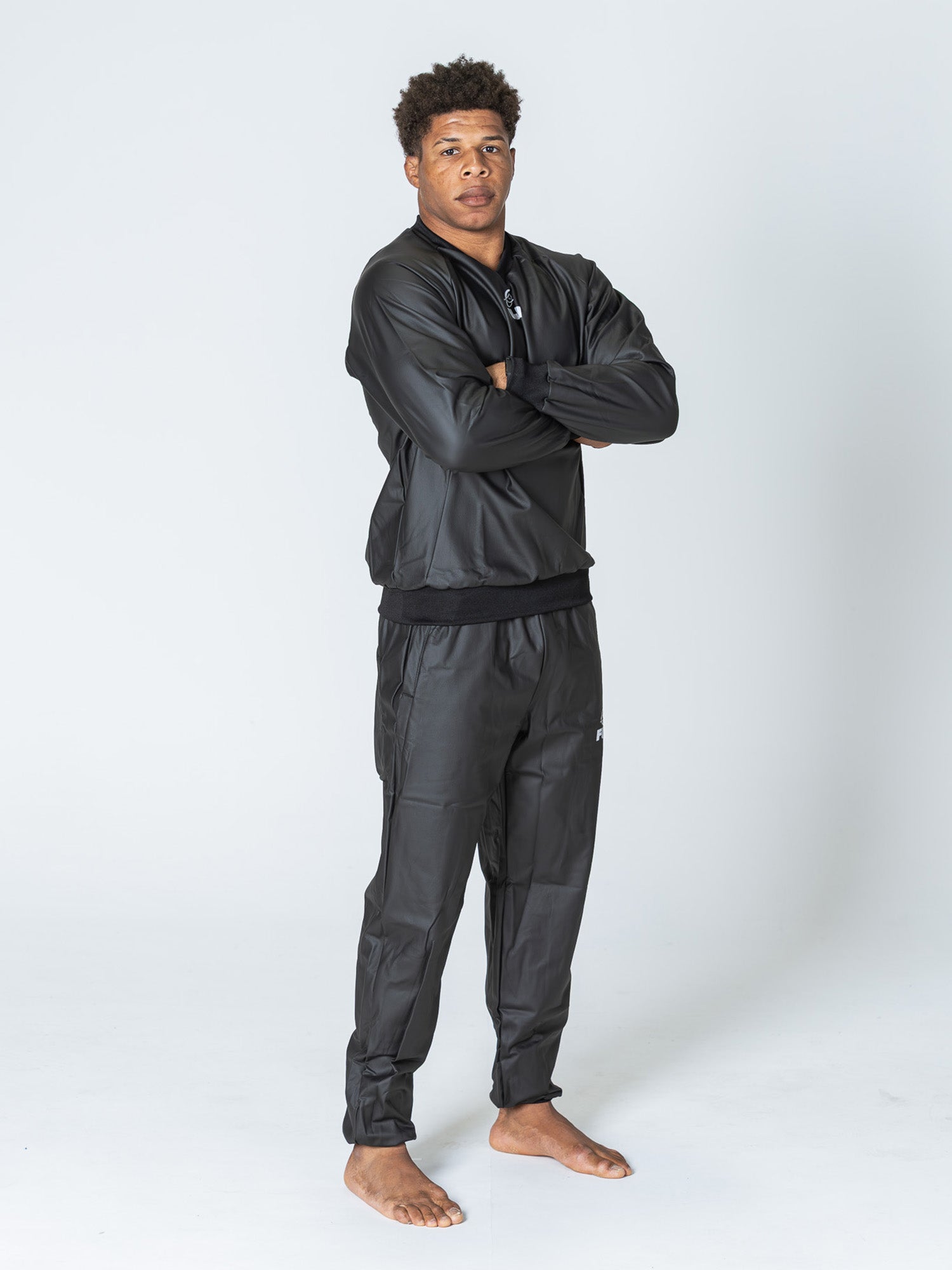 A man stands barefoot with arms crossed, wearing the Grappling Sauna Suit Black, against a plain white background.