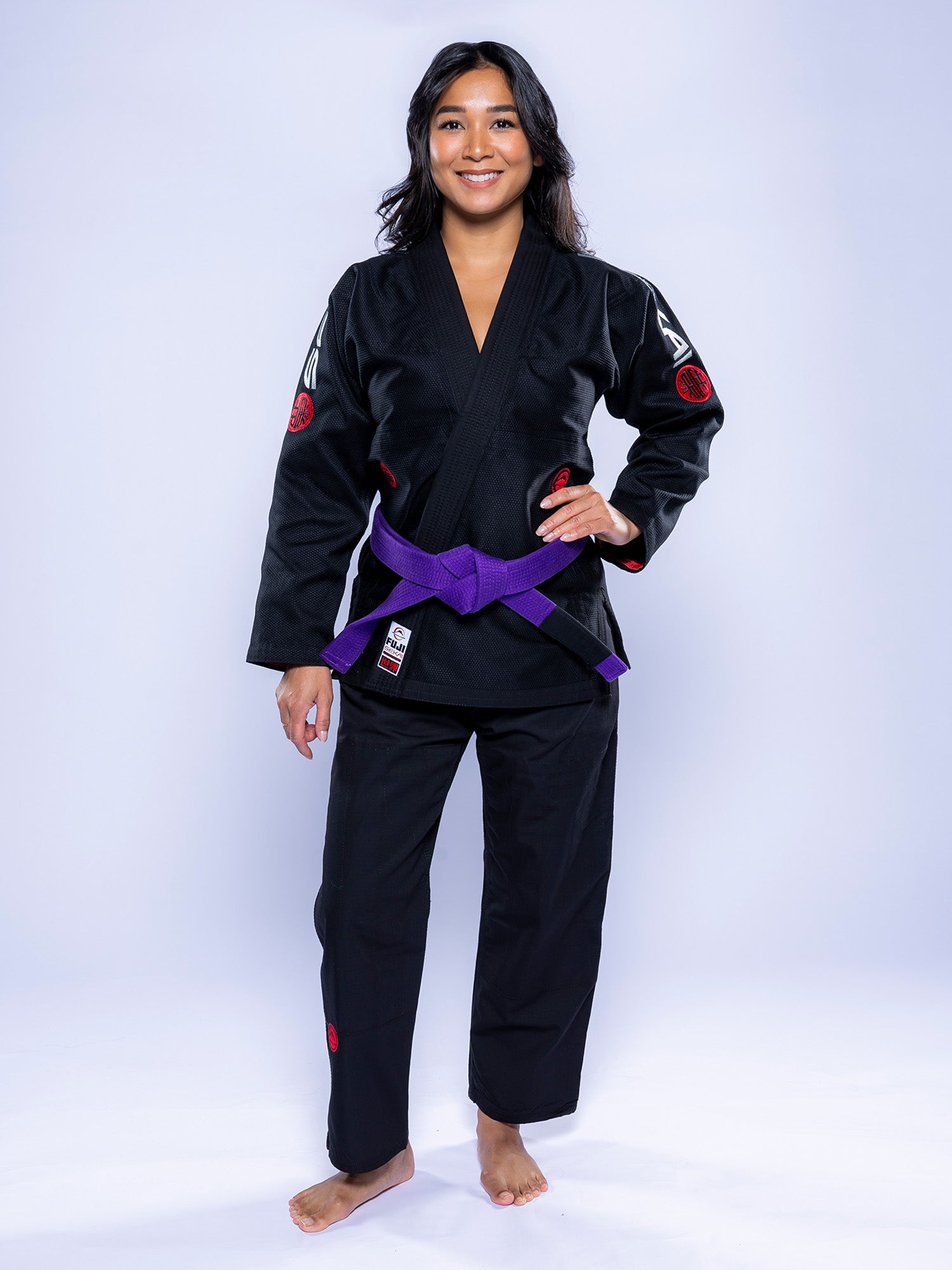 A woman stands barefoot on a white background, smiling and wearing the Womens SEKAI 3.0 BJJ Gi Black Grey with a purple belt tied at her waist, her hands resting at her sides.