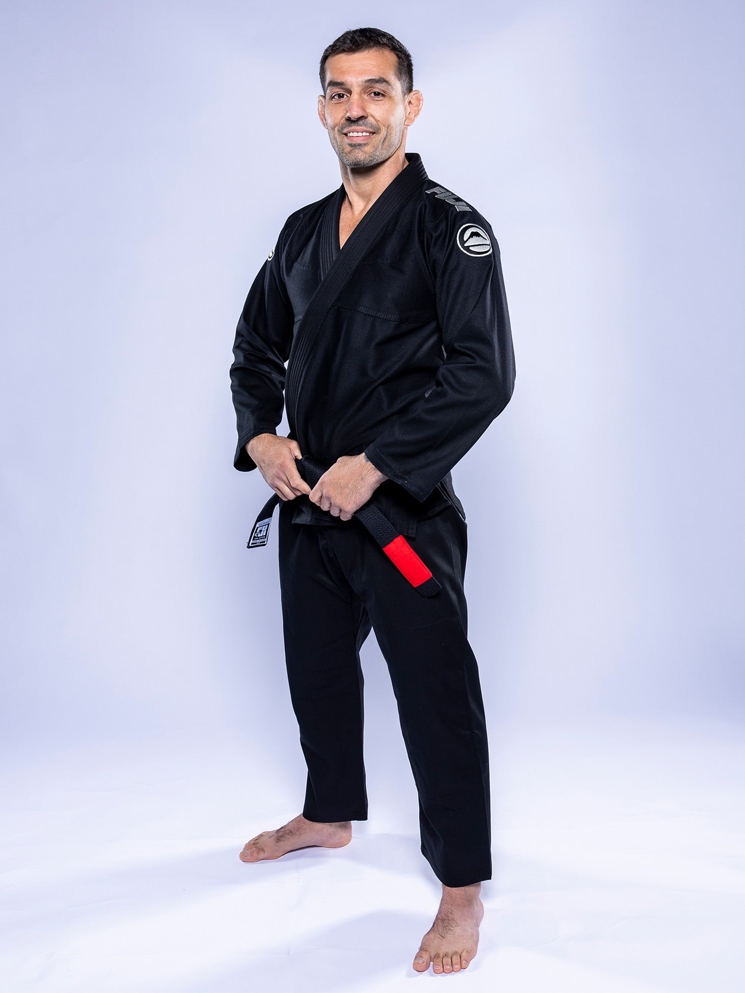A man stands barefoot on a white floor, smiling confidently in the Baseline 2.0 BJJ Gi Black Grey with a red and black belt, holding the belt’s ends against a plain light background.