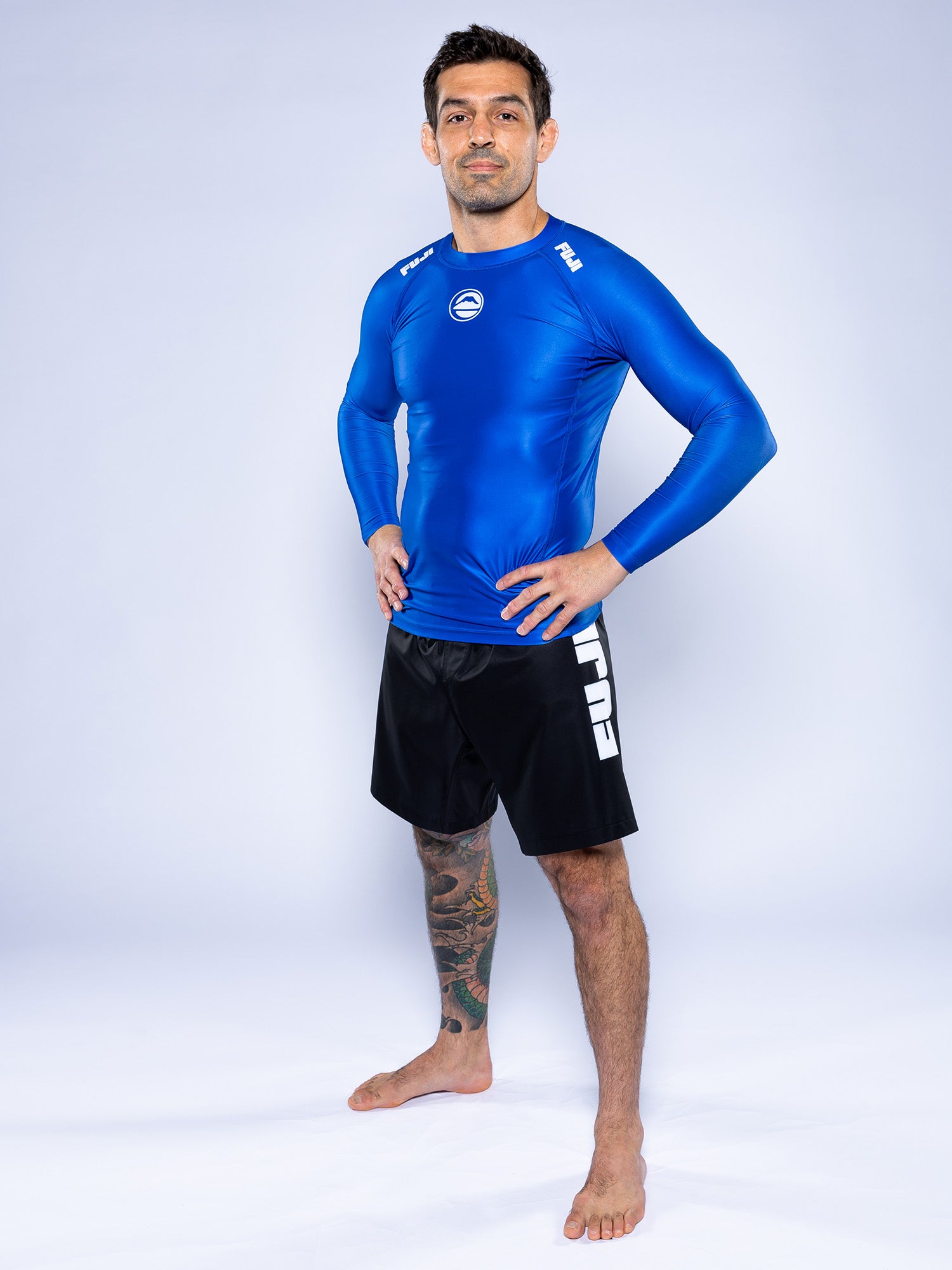 A man stands barefoot on a white background, wearing the Baseline 2.0 Long Sleeve Rashguard Blue and black shorts with FUJI branding. He has tattoos on his left leg and rests his hands on his hips.