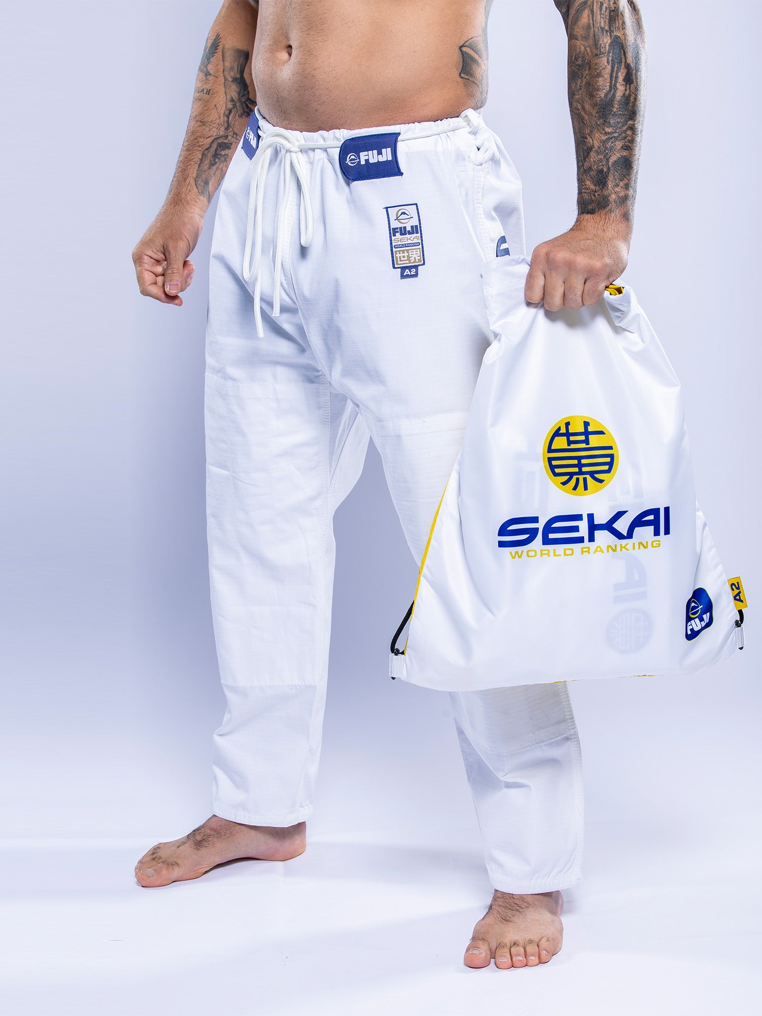 A person in a SEKAI 3.0 BJJ Gi White Navy holds a matching white drawstring bag labeled SEKAI WORLD RANKING with blue and yellow logos, standing barefoot against a light background.