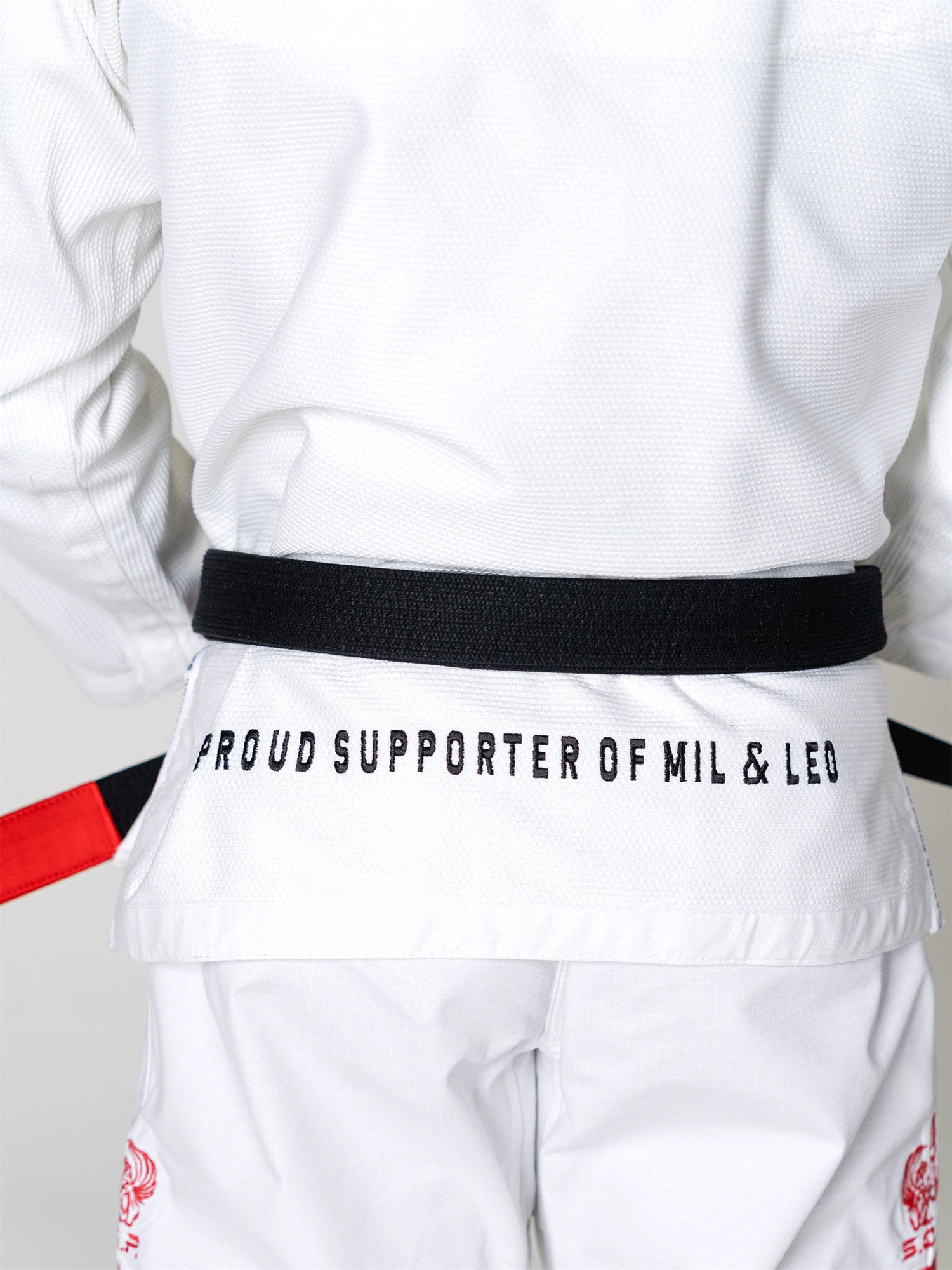 Someone wearing the S.O.C.P. Sekai BJJ Gi 2.0 White and a black belt is seen from the back; "PROUD SUPPORTER OF MIL & LEO" is printed in black on the jacket.