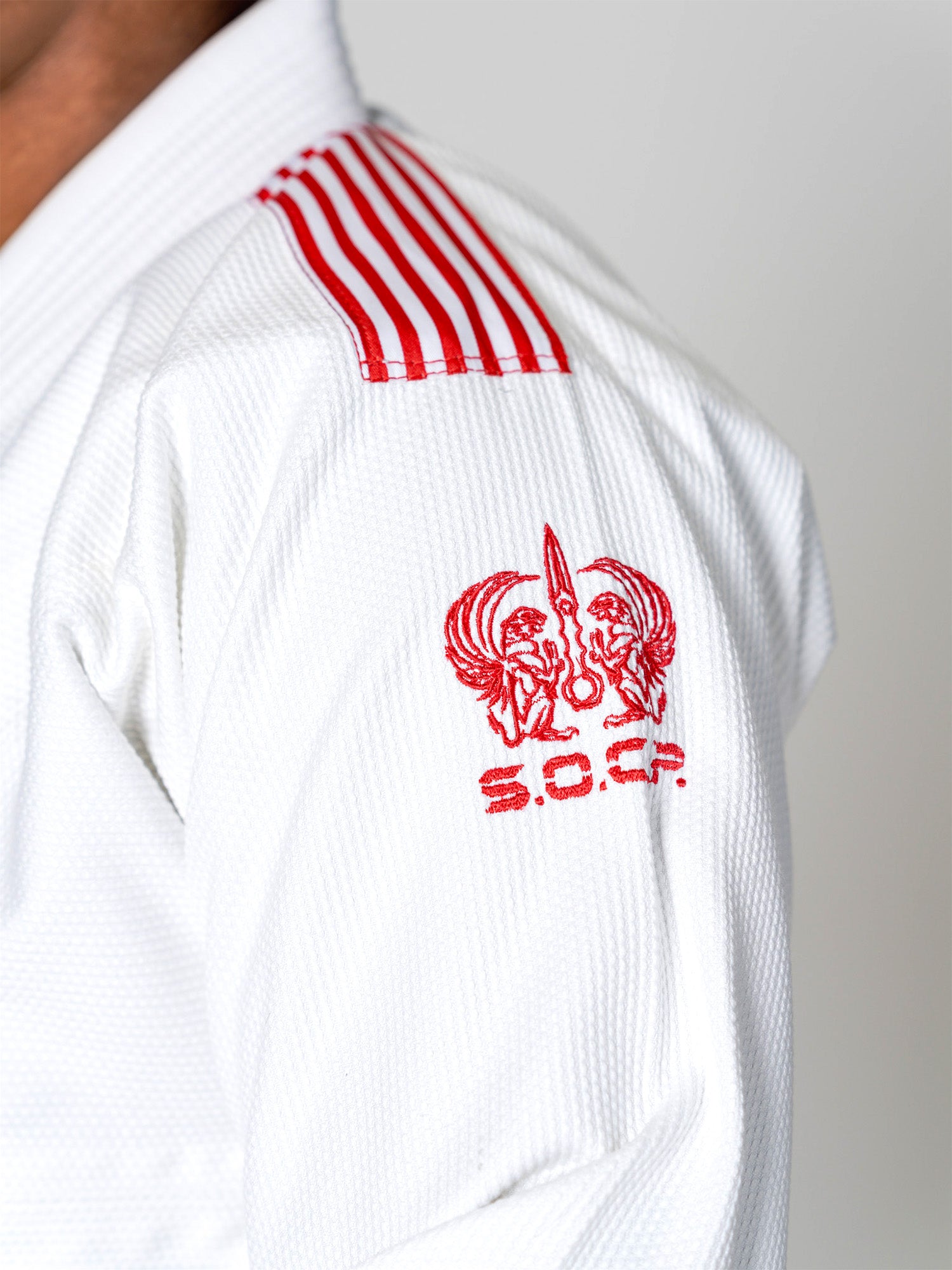 Close-up of a person wearing the S.O.C.P. Sekai BJJ Gi 2.0 White with red shoulder stripes and an eagle-embroidered S.O.C.P. emblem on the sleeve.