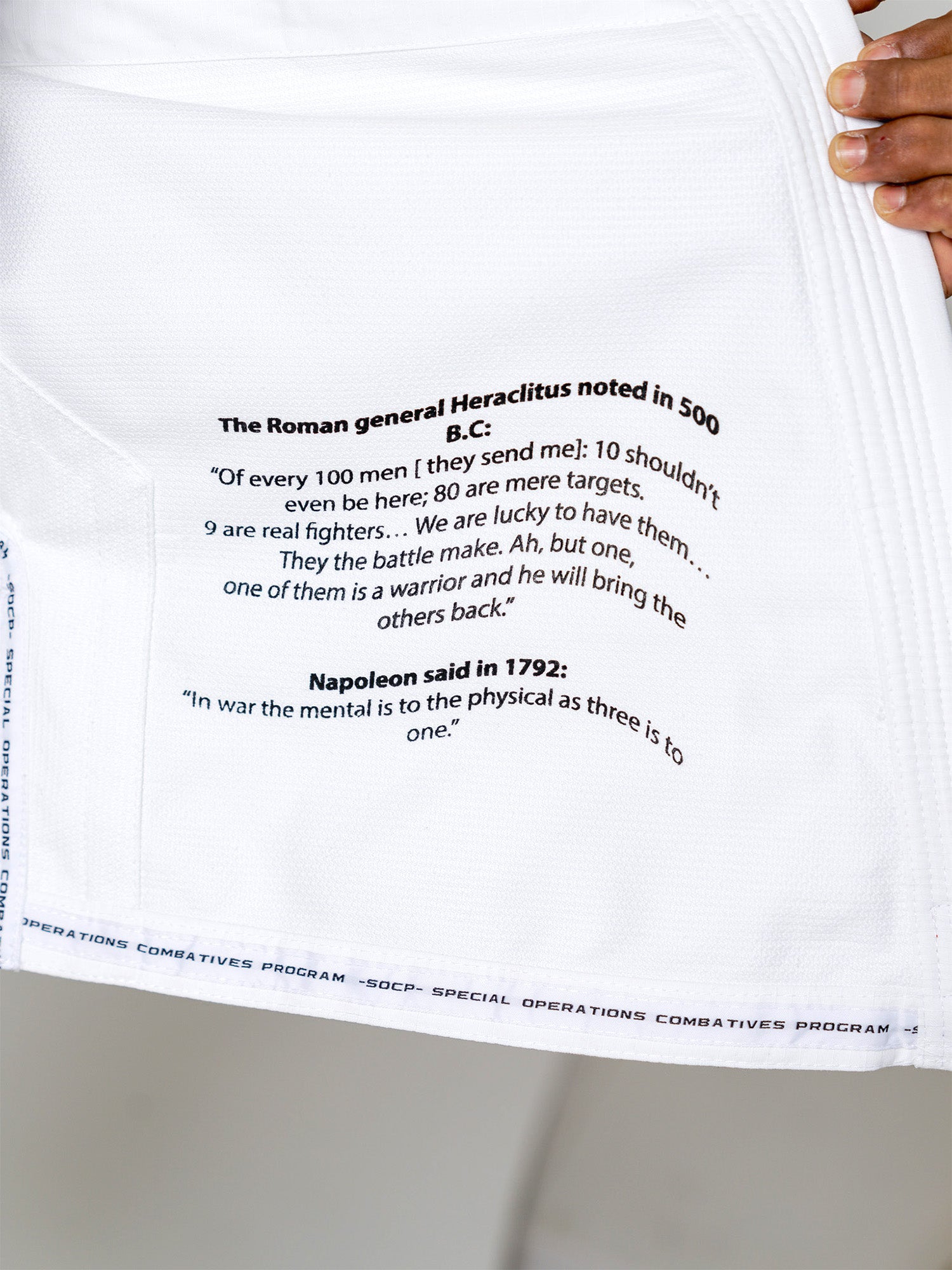 A close-up of the S.O.C.P. Sekai BJJ Gi 2.0 White, featuring printed quotes about war and warriors attributed to Heraclitus and Napoleon. A hand holds open the fabric to display the text.