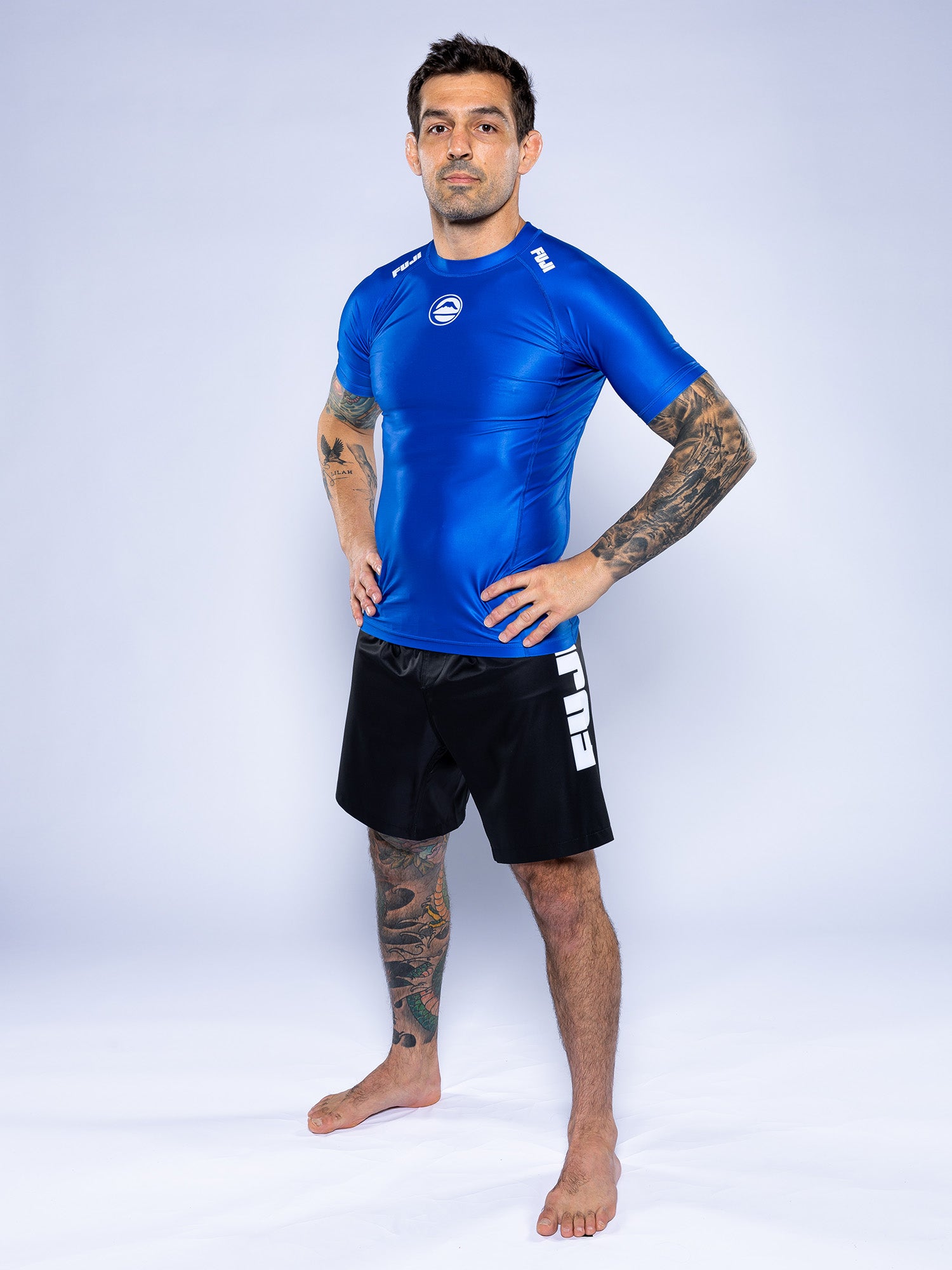 A tattooed man stands barefoot on a white background, confidently posing with hands on hips while wearing the Baseline 2.0 Short Sleeve Rashguard Blue and black shorts with “FUJI” in white on the left leg.