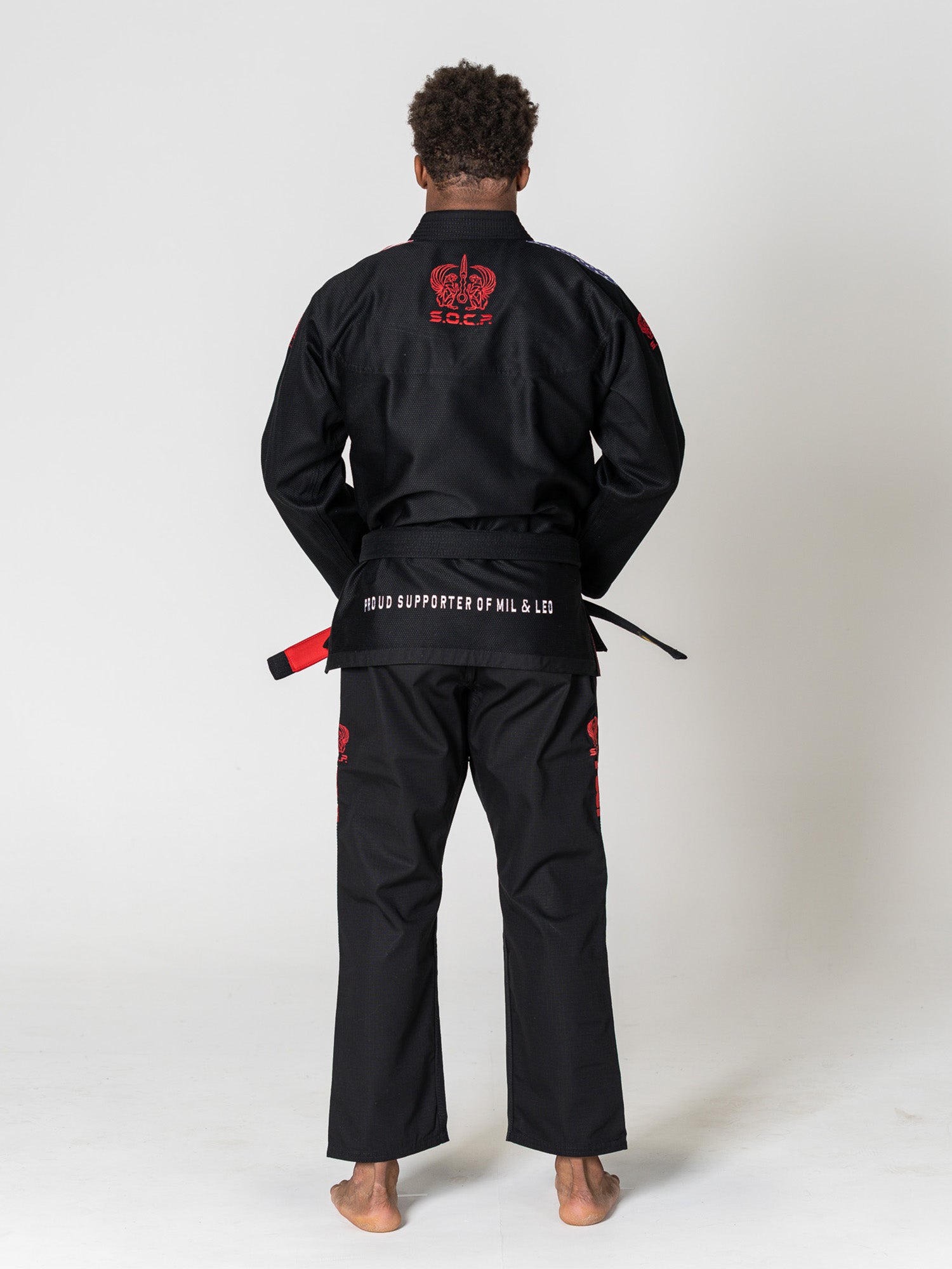 A person stands barefoot, facing away, wearing the S.O.C.P. Sekai BJJ Gi 2.0 Black with red designs and a black belt. The back of the jacket features a crown graphic and text. The background is plain white.
