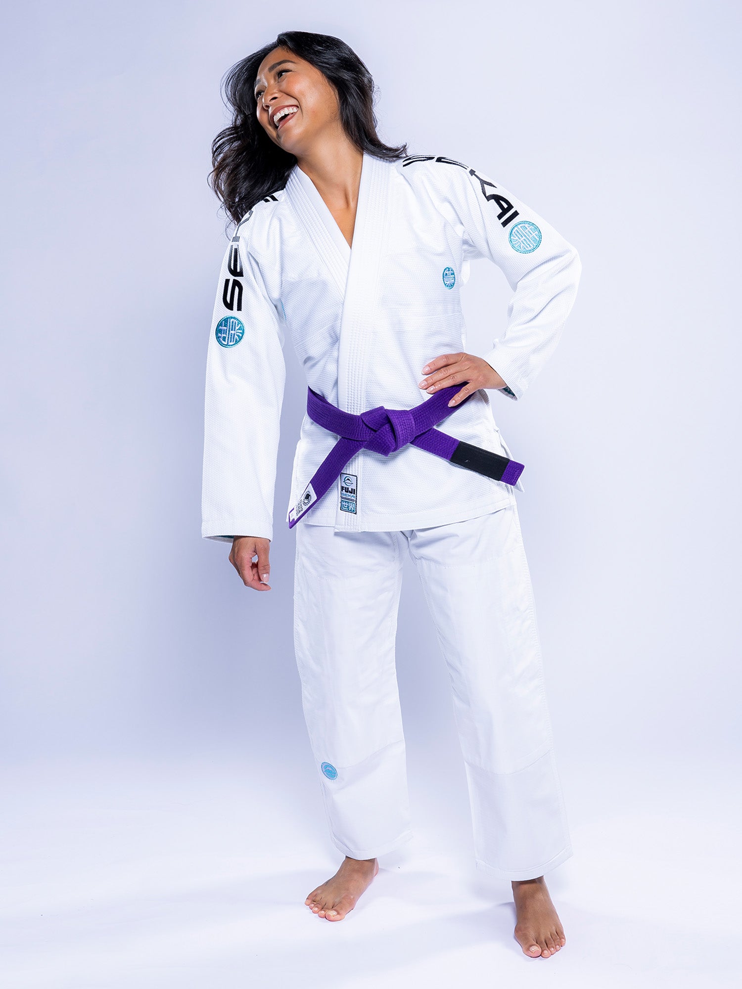 A woman wearing the Womens SEKAI 3.0 BJJ Gi White Teal with a purple belt stands barefoot, smiling and looking to the side against a plain white background.