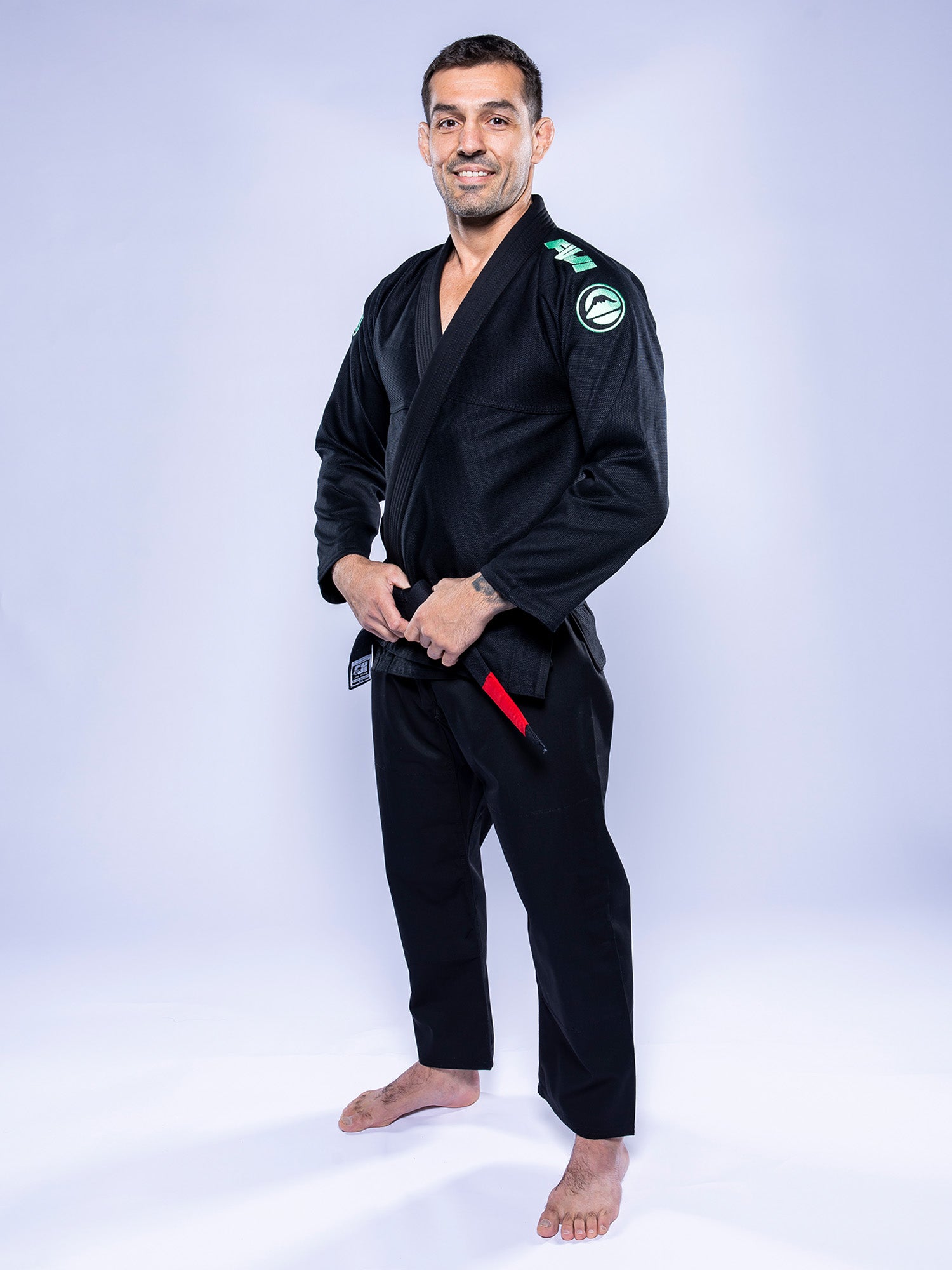 A man wearing the Baseline 2.0 BJJ Gi Black Mint and a black belt stands barefoot on a white surface, smiling at the camera against a light background.