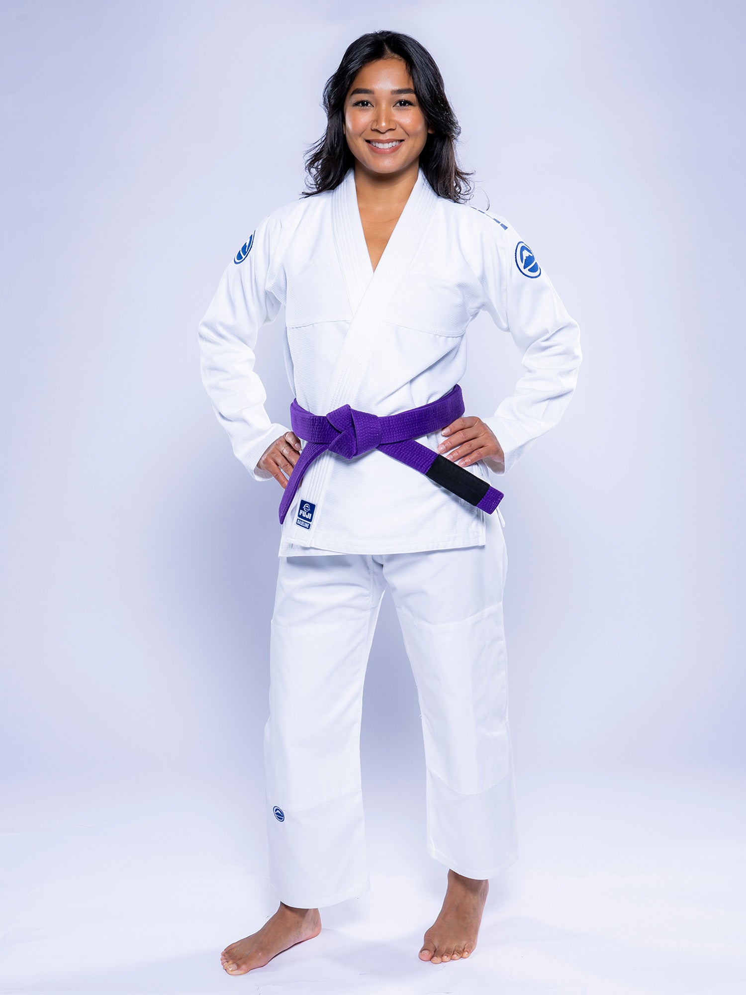 A woman stands barefoot on a white background, smiling with her hands on her hips while wearing the Womens Baseline 2.0 BJJ Gi White Navy and a purple belt.