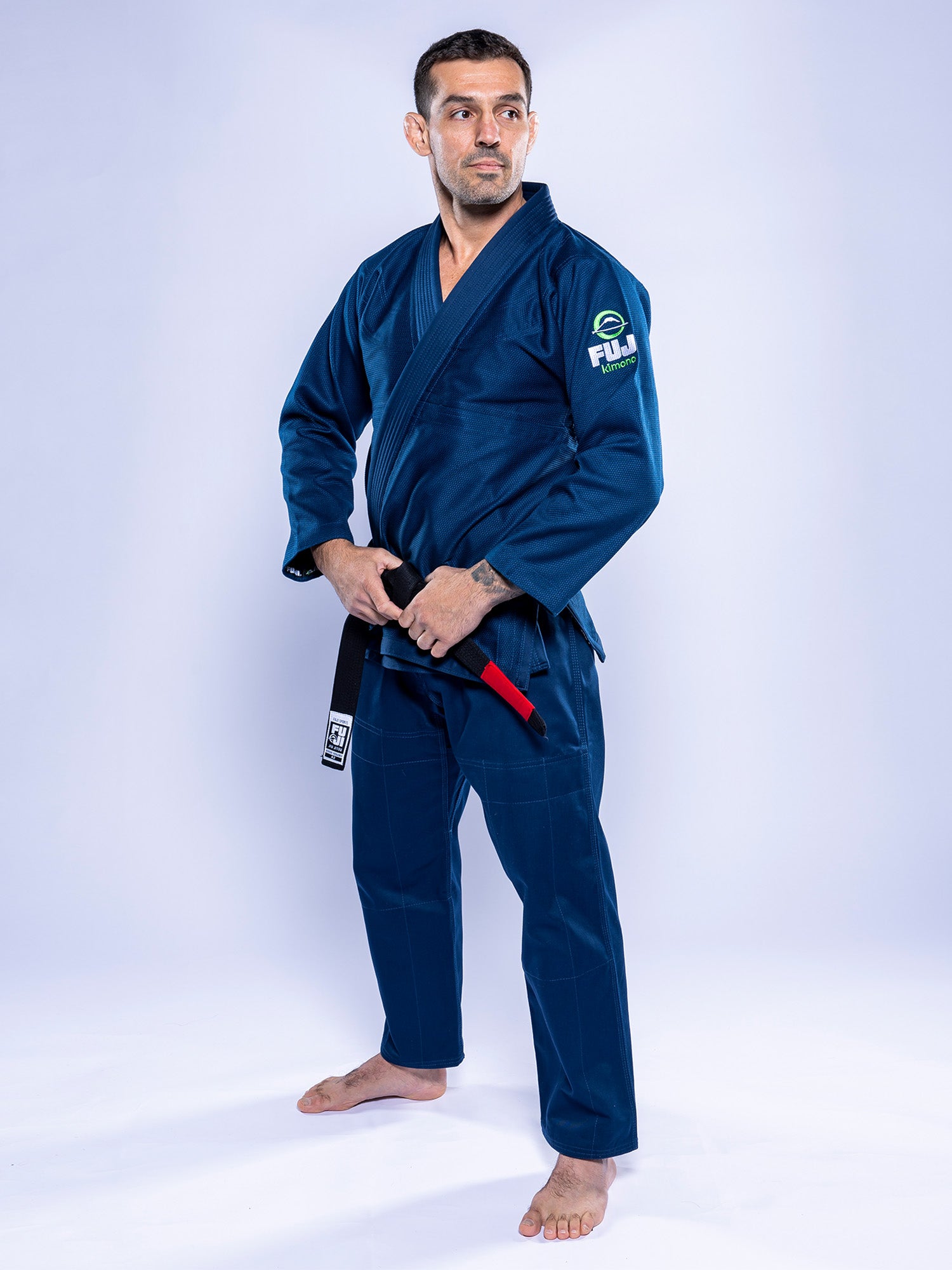 A man wearing the Lightweight BJJ Gi Navy and a black belt stands barefoot on a white background, turned slightly to the side with his hands holding his belt.