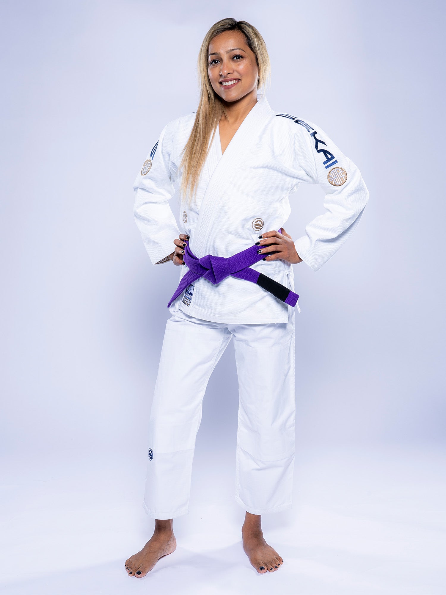 A woman stands confidently, smiling with hands on hips, wearing the Womens SEKAI 3.0 BJJ Gi White Navy and a purple belt against a plain light background.