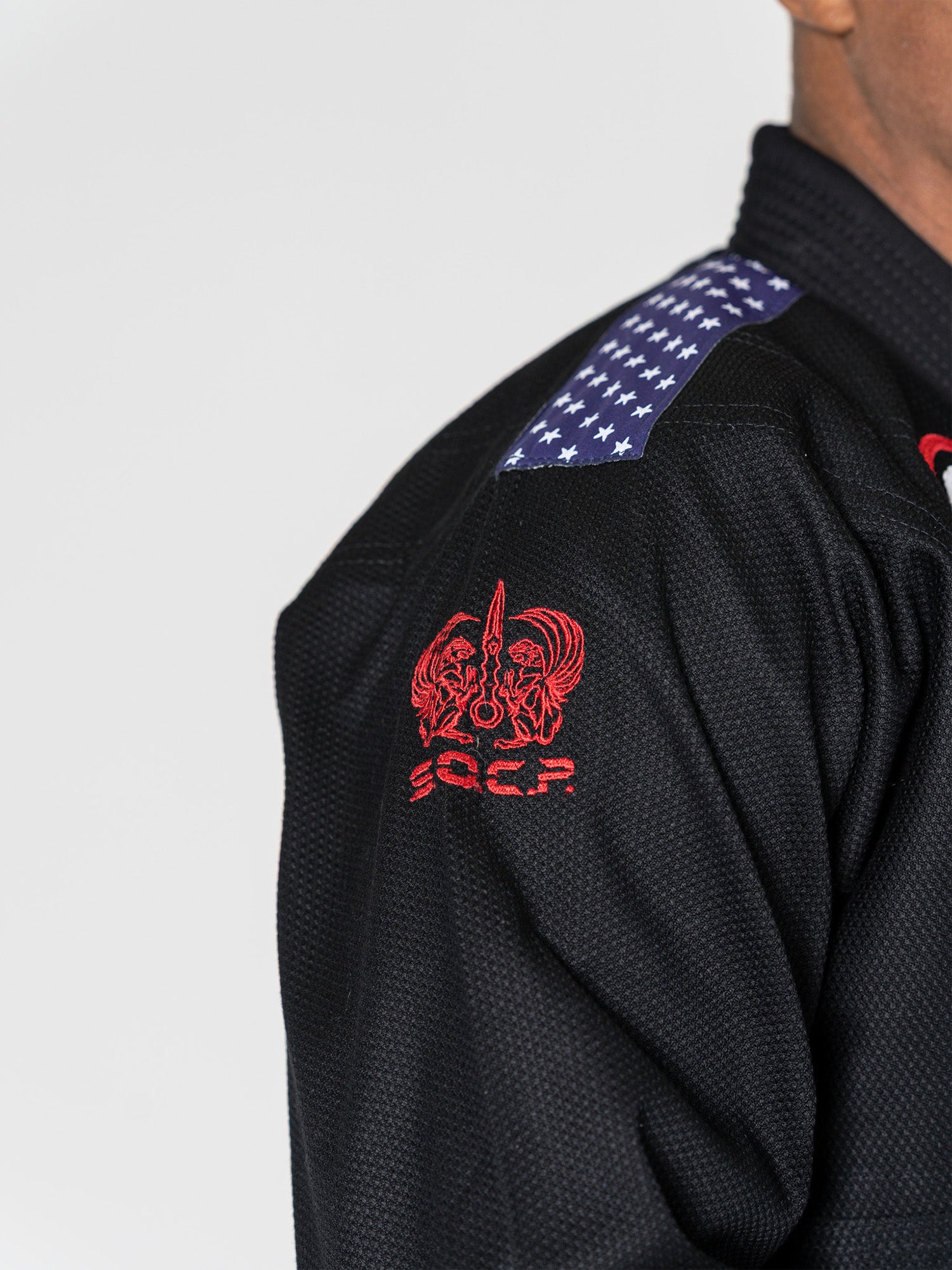 Close-up of a person wearing the S.O.C.P. Sekai BJJ Gi 2.0 Black, featuring a patch of white stars on a dark shoulder background and red embroidered design and letters on the upper arm.