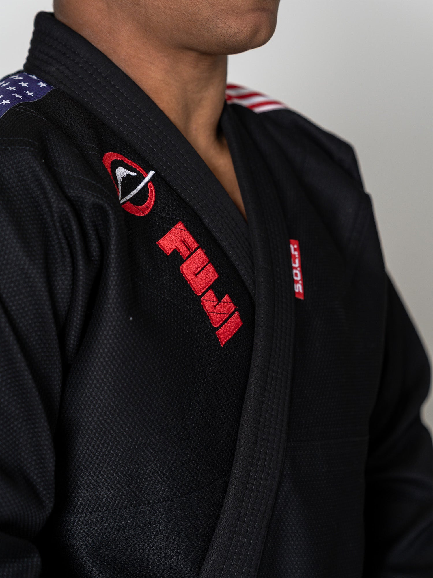 A person wearing the S.O.C.P. Sekai BJJ Gi 2.0 Black, featuring red and white embroidery and an American flag patch on the shoulder. Only their upper body and part of their face are visible against a plain background.