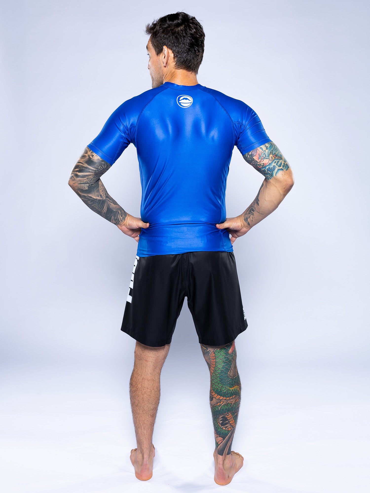 A man with tattoos on his arms and legs stands barefoot, hands on hips, facing away in a Baseline 2.0 Short Sleeve Rashguard Blue and black shorts against a plain light background.