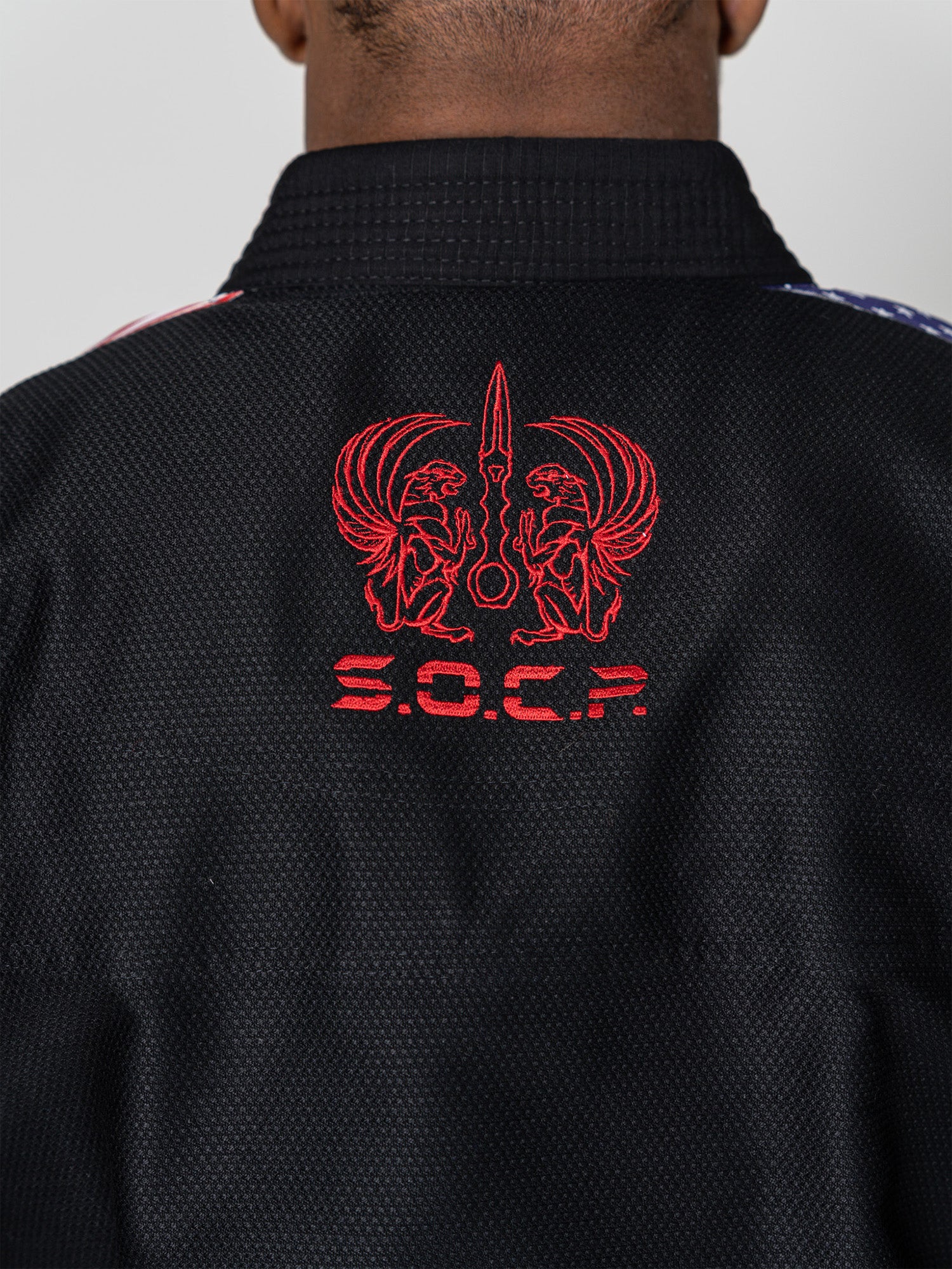 A person wearing the S.O.C.P. Sekai BJJ Gi 2.0 Black, featuring a red embroidered design on the back with two winged figures, a stylized sword, and the letters S.O.C.P. below.