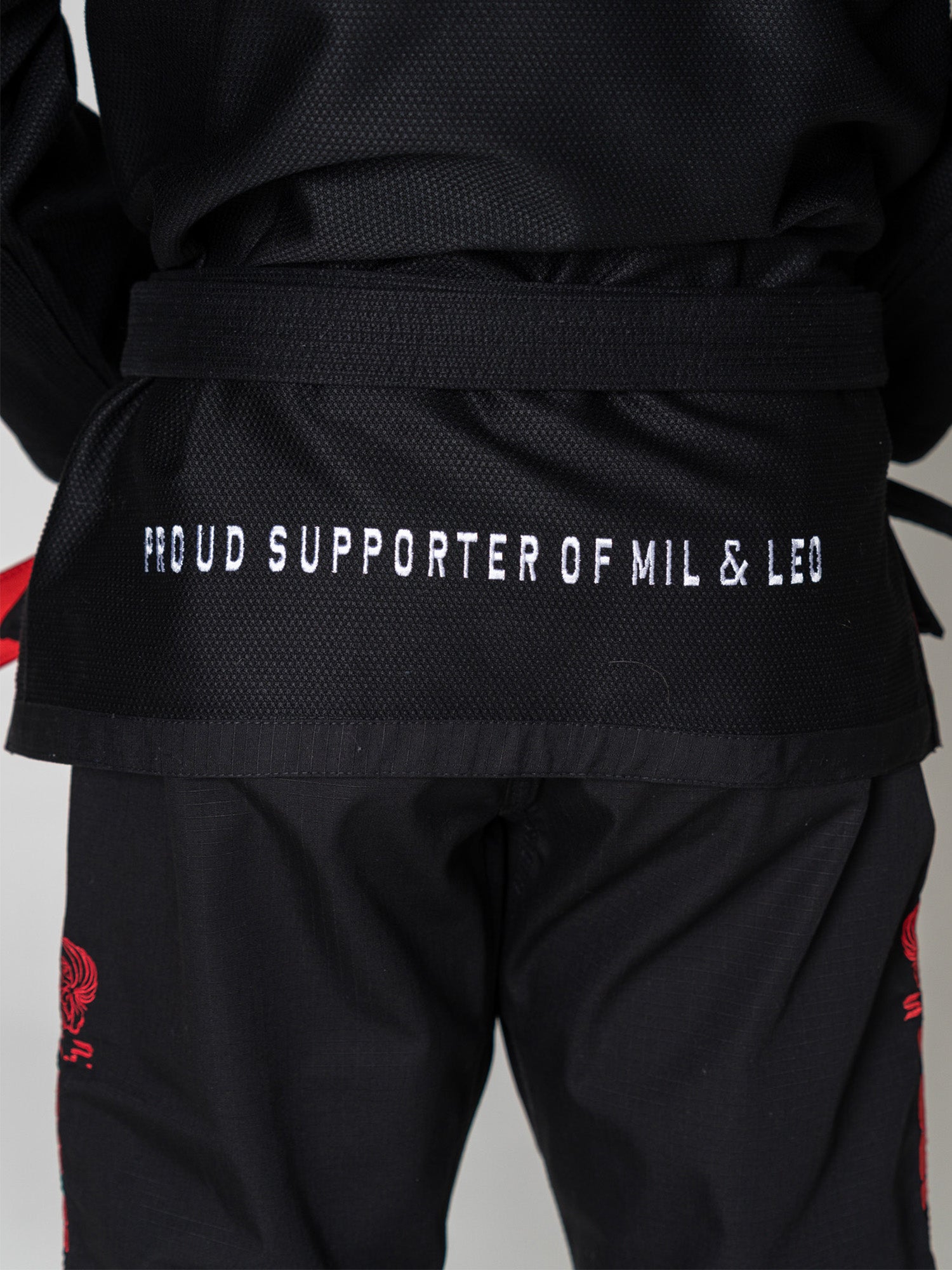 A person wearing the S.O.C.P. Sekai BJJ Gi 2.0 Black stands with hands behind their back, displaying white text on the jacket that reads "PROUD SUPPORTER OF MIL & LEO.