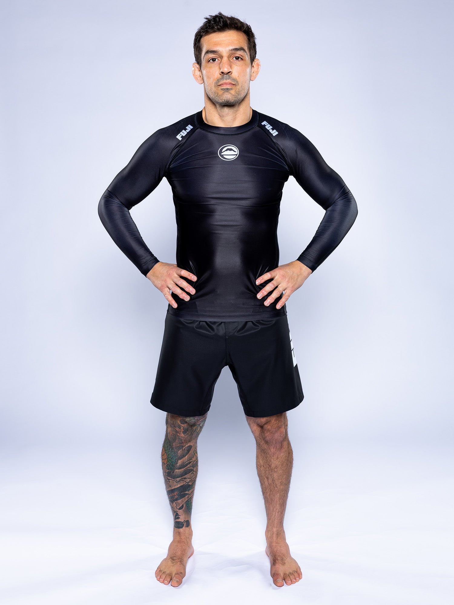 A man stands barefoot on a white surface, wearing the Baseline 2.0 Long Sleeve Rashguard Black and black shorts. He has a tattoo on his left leg, with hands on hips against a plain, light background.