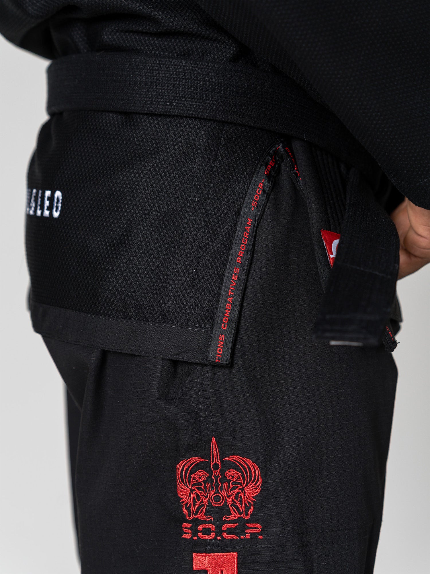 A close-up shows someone wearing the S.O.C.P. Sekai BJJ Gi 2.0 Black with a black belt. Red embroidered designs and text appear on the jacket and pants as their hand rests near their waist.