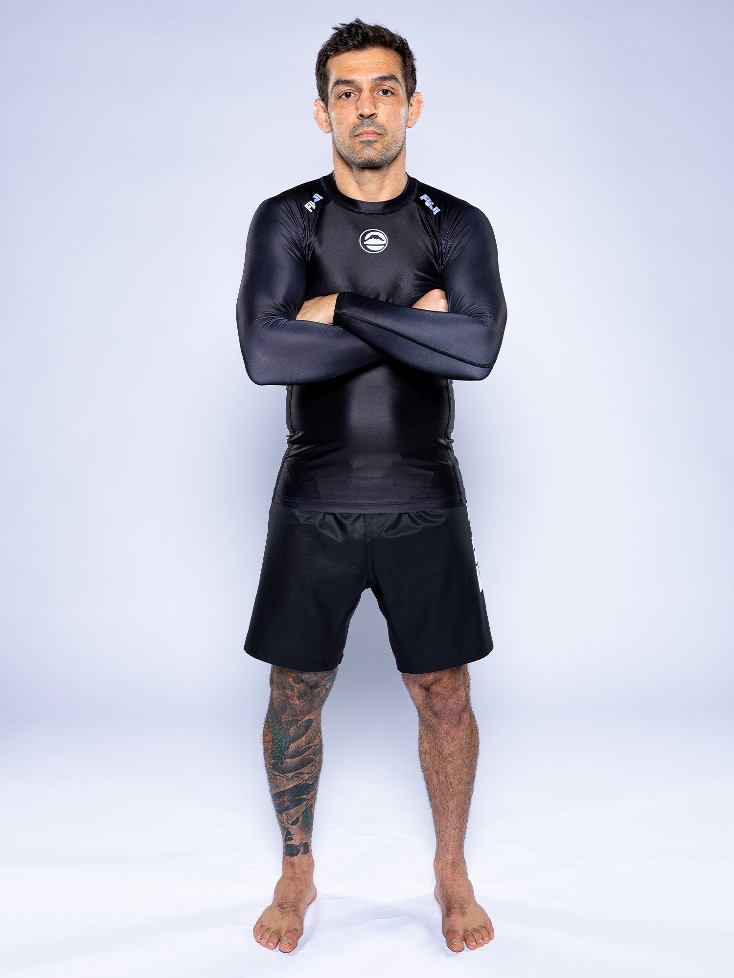 A man with short dark hair and a large tattoo on his left leg stands barefoot against a light background, arms crossed, wearing the Baseline 2.0 Long Sleeve Rashguard Black and matching black shorts, with a neutral expression.