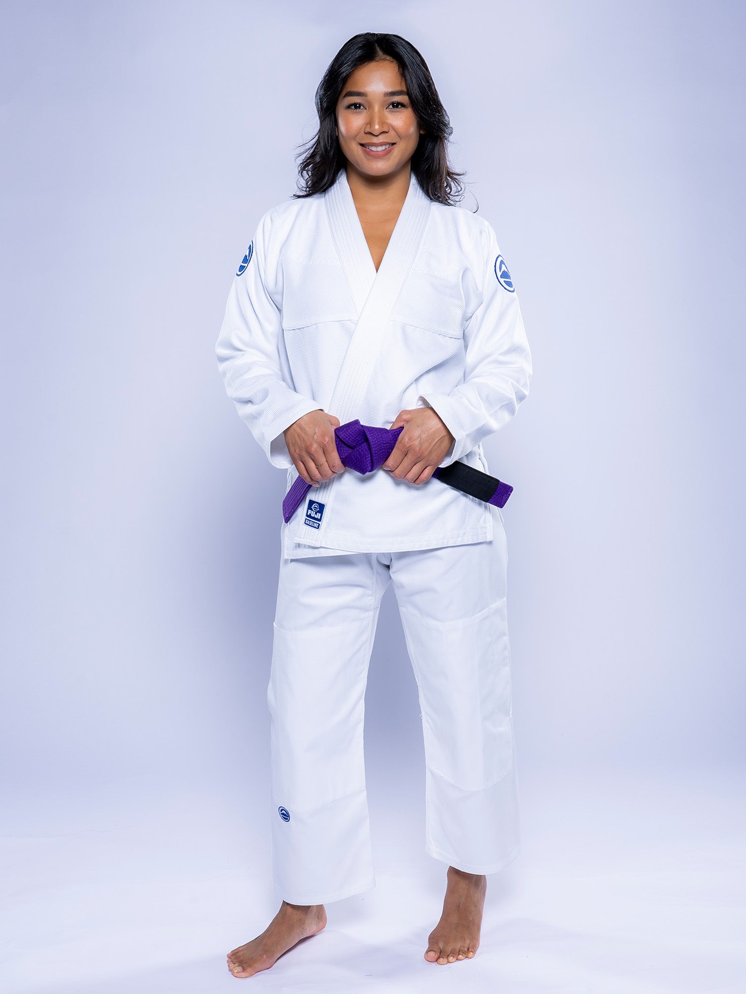 A woman stands barefoot on a white background, smiling and wearing the Womens Baseline 2.0 BJJ Gi White Navy with a purple belt. Her hands rest on the belt, and her hair is loose.