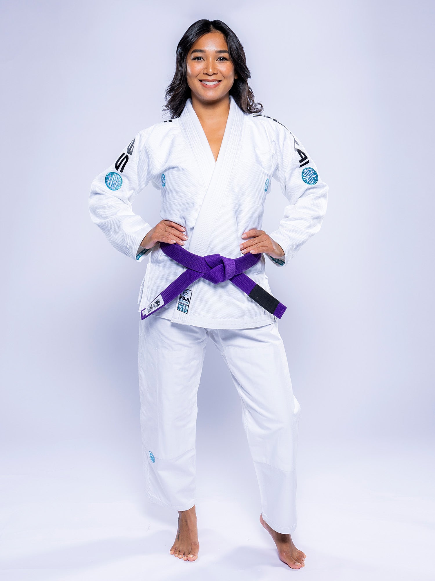 A woman stands barefoot on a white background, smiling confidently with her hands on her hips while wearing the Womens SEKAI 3.0 BJJ Gi White Teal and a purple belt.