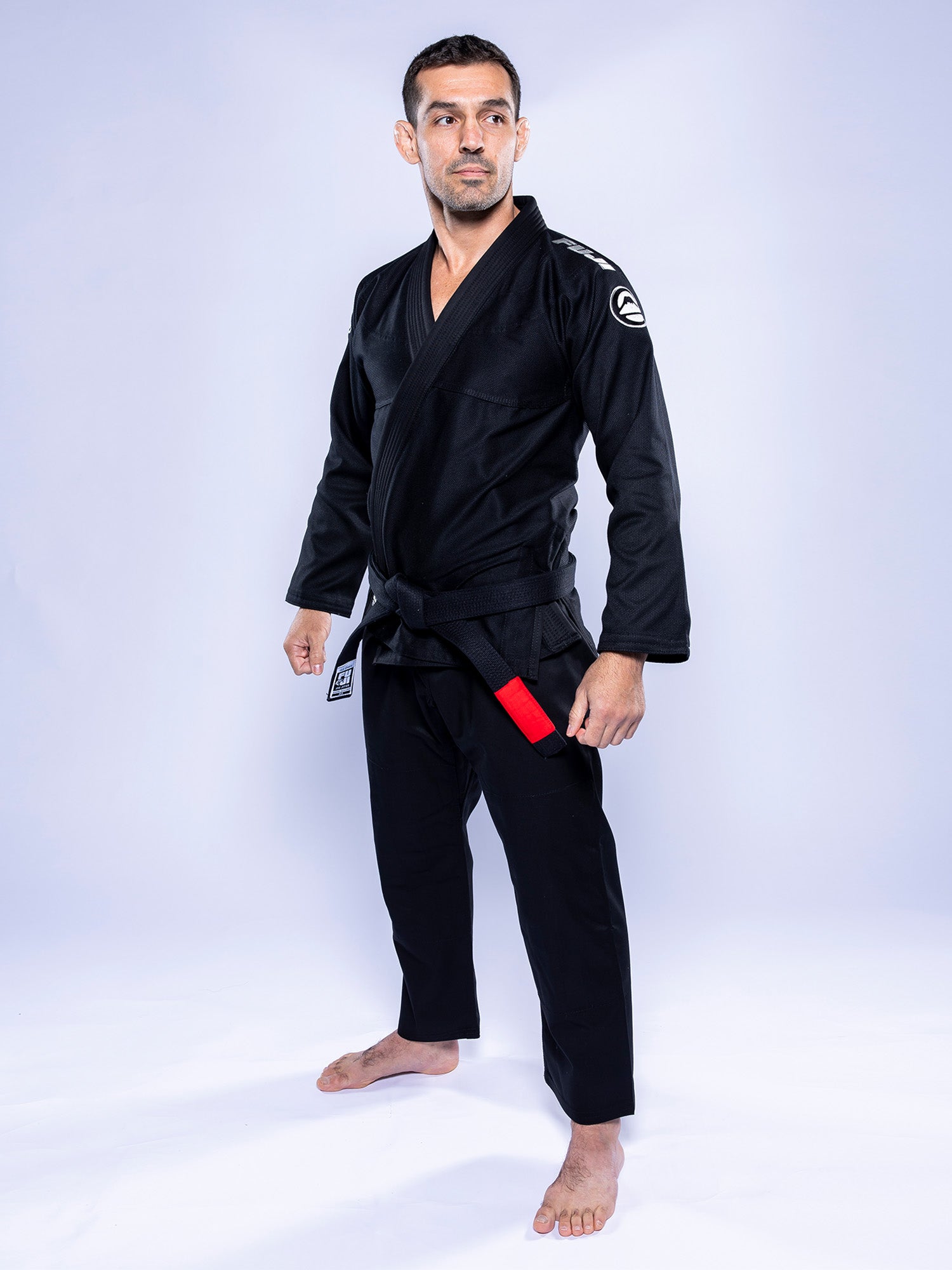 A man in the Baseline 2.0 BJJ Gi Black Grey stands barefoot on a white background, looking to the side with a serious expression. The gi features a shoulder patch and a red tag on the belt.